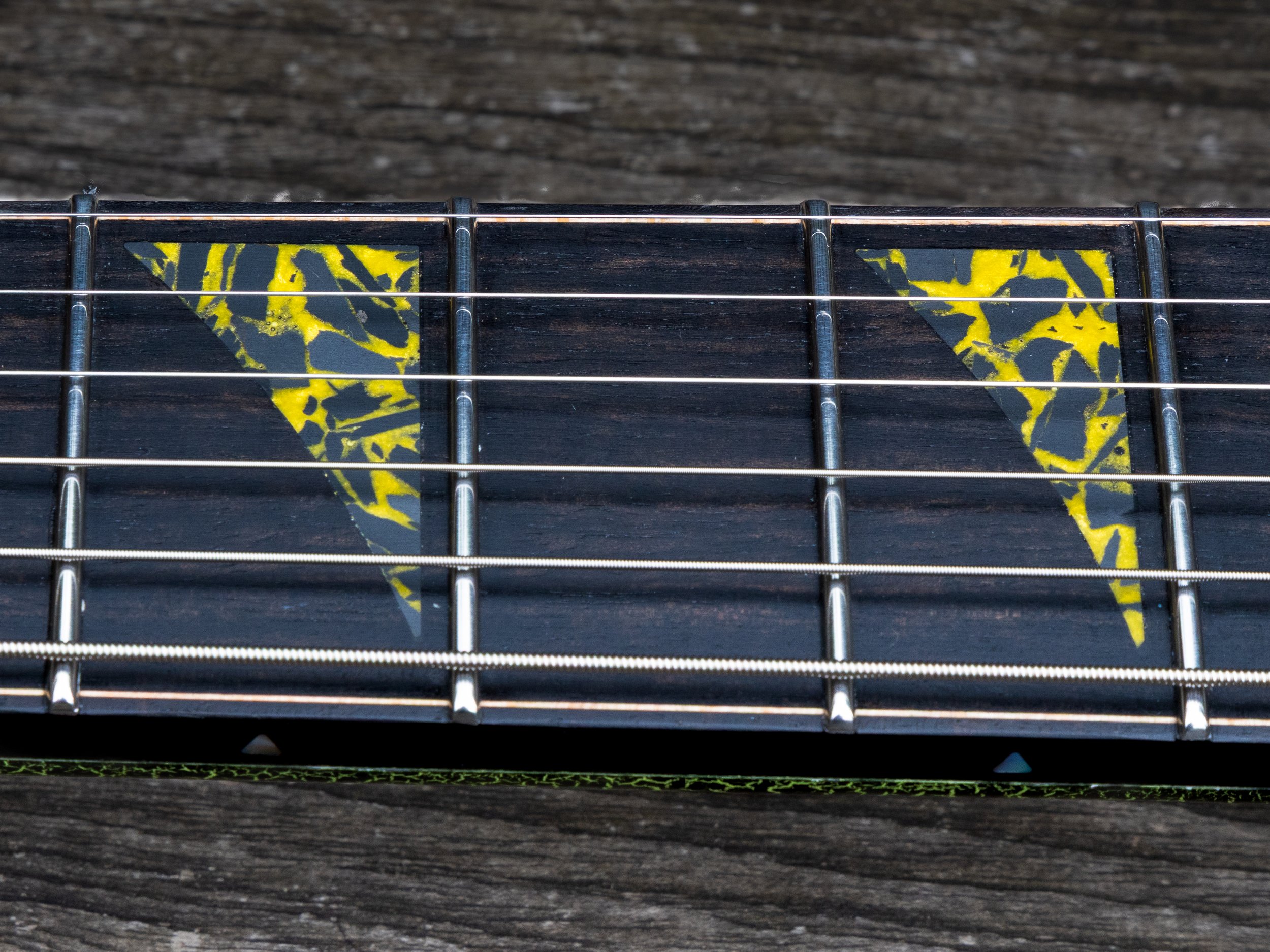 Close-up of the fretboard of an acoustic guitar showing some black and yellow patterned inlays and metal frets on a wooden surface.
