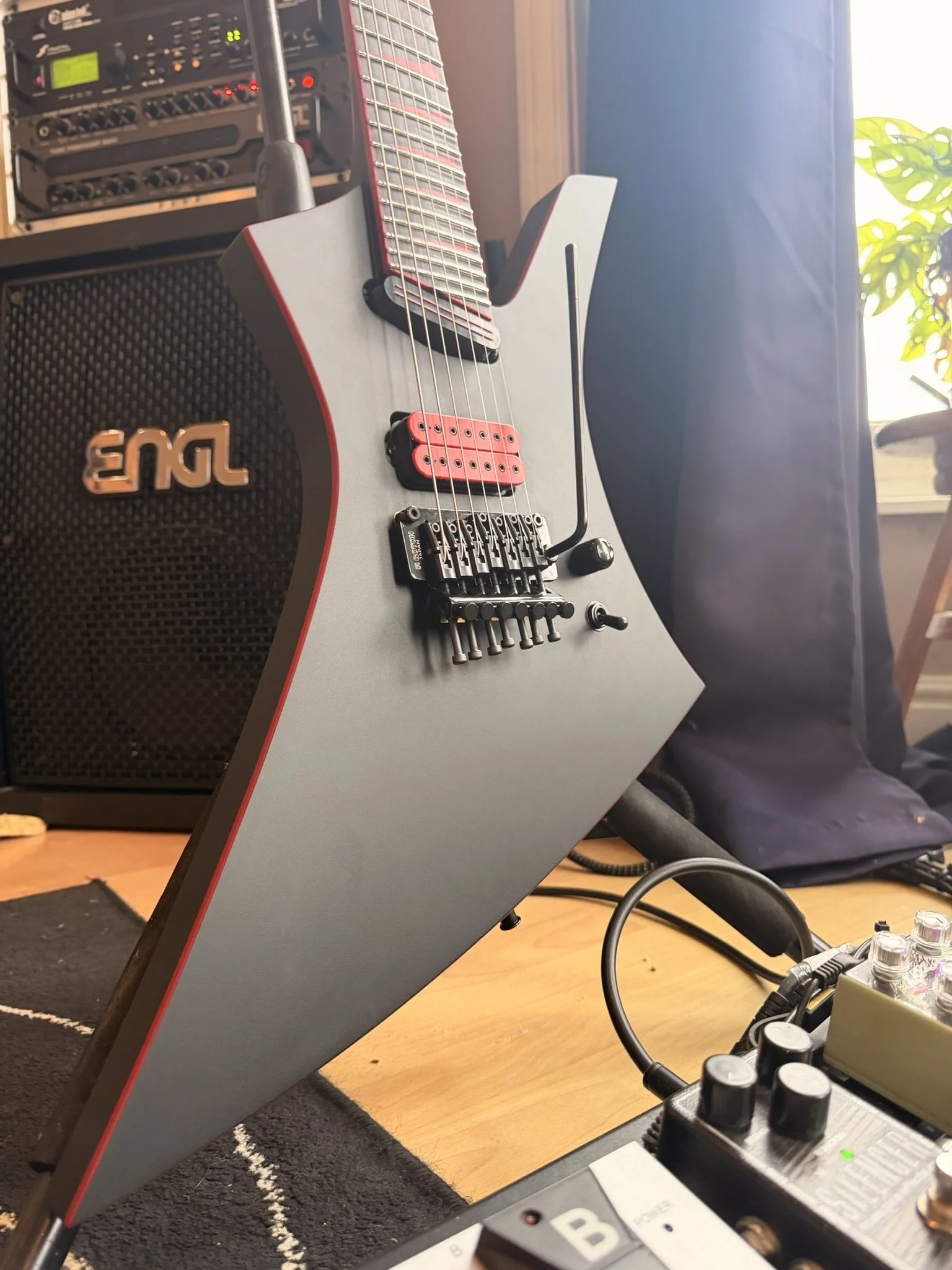 An electric guitar with a unique angular body design sitting on a floor next to a guitar amplifier, with music equipment and cables nearby, in a room with wooden flooring and a nearby window with natural light.