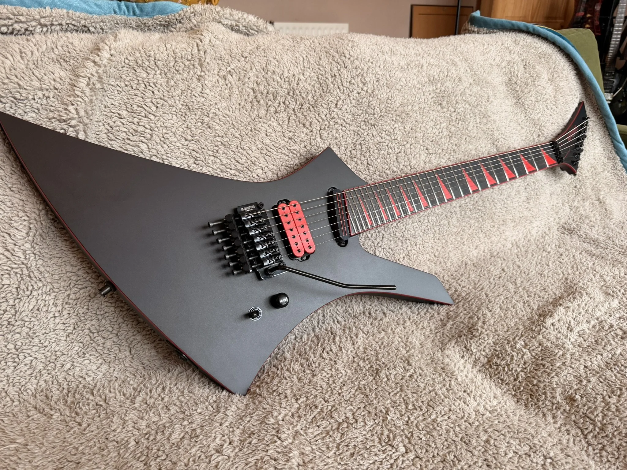 An electric guitar with a black body, red accents, and a unique angular shape is lying on a beige, fluffy carpet. The guitar has a black tremolo arm and a dual humbucker pickup system, with red pickups and a matching red bridge.