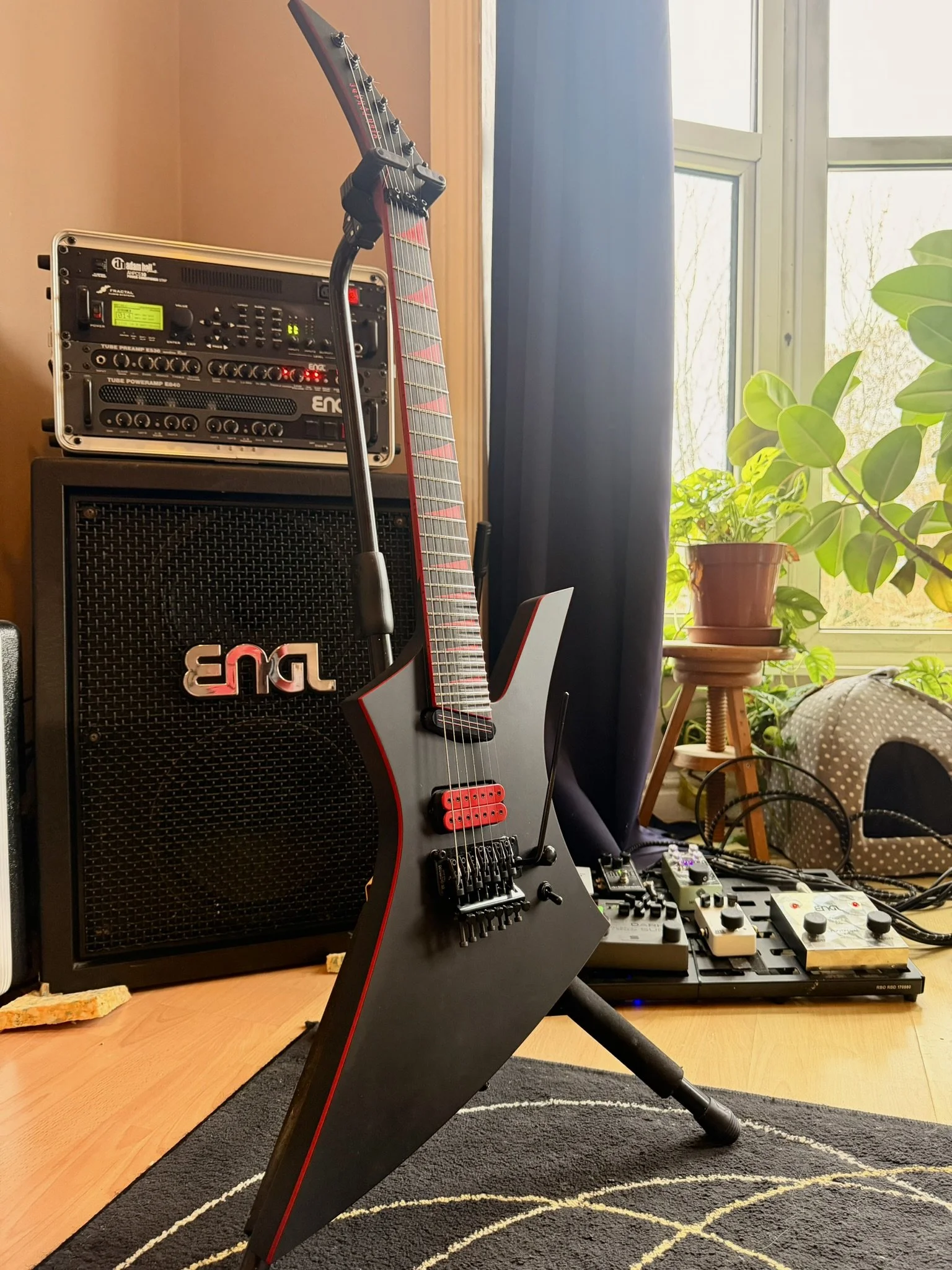 A black and red electric guitar with a stand is in front of music equipment, including a black amplifier and audio gear. The setup is in a room with wooden floors, a black rug with white patterns, and a window with blue curtains, sunlight, and green 