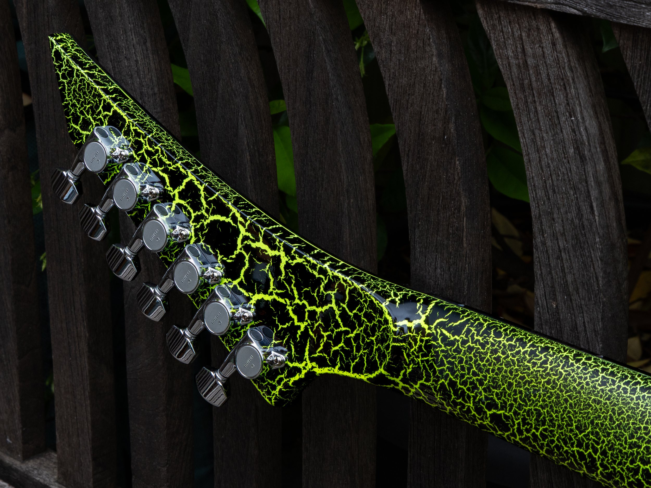 Close-up of a guitar headstock with black tuning pegs and a neon green lightning pattern on the fretboard, resting against a dark wooden surface with green leaves in the background.