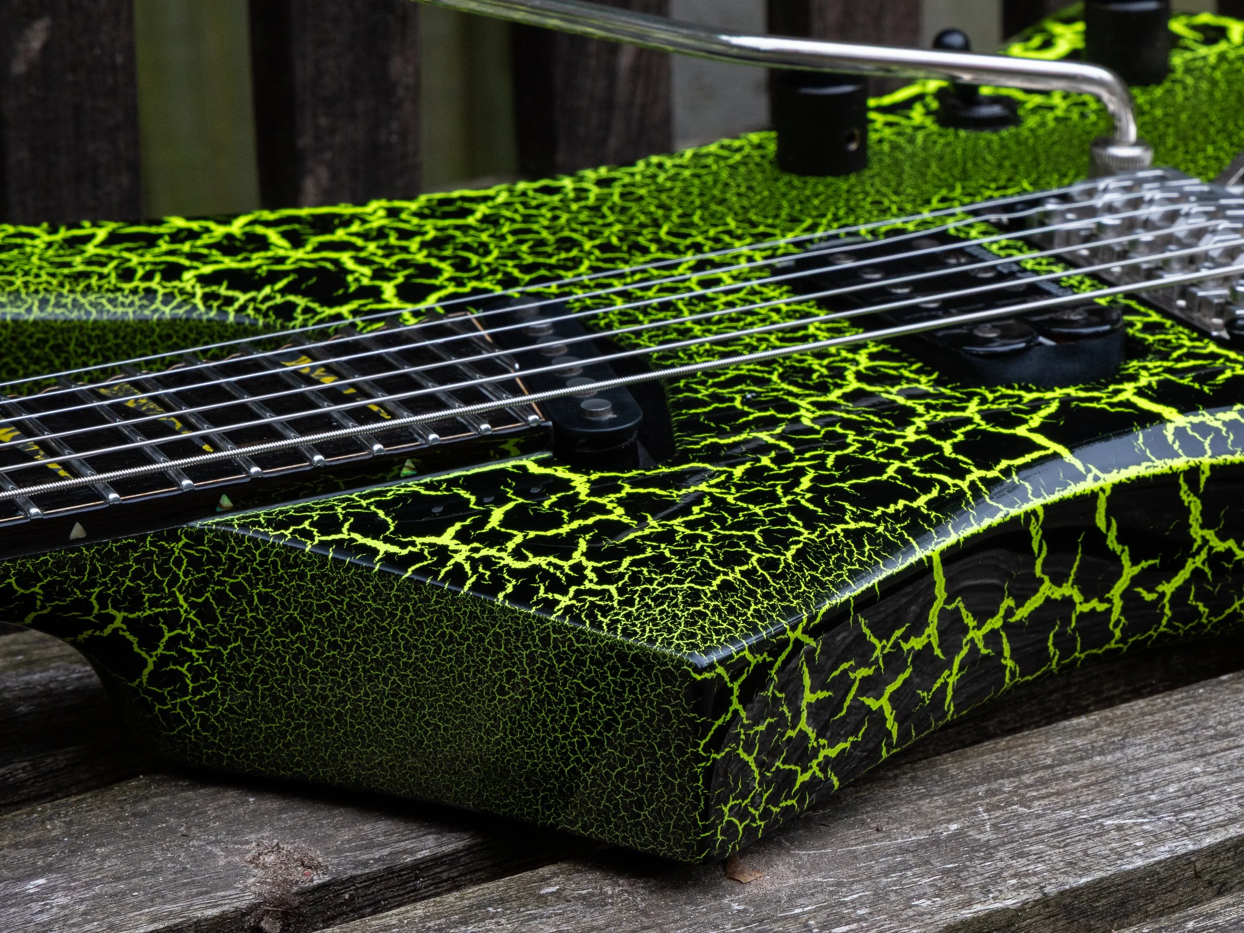 Close-up of a guitar with a black and neon green cracked pattern finish, metal strings, and tuning pegs on a wooden surface.
