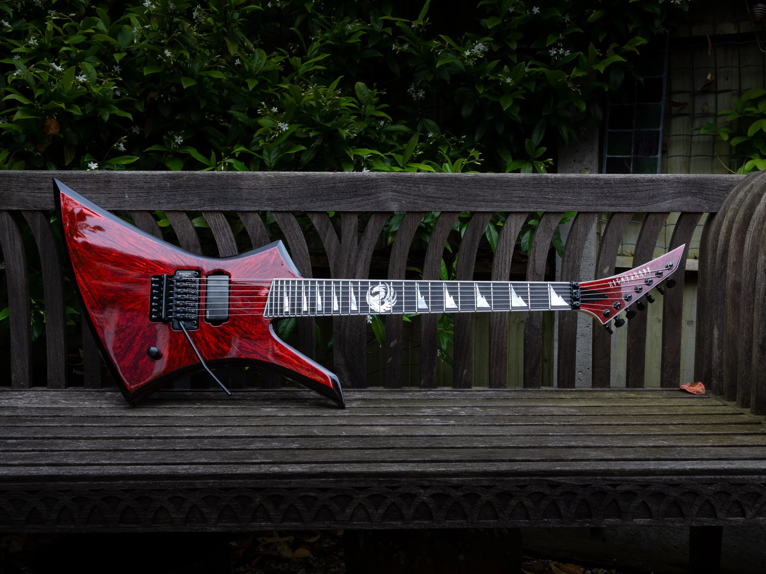A red electric guitar resting on a wooden garden bench on a patio, with green leafy bushes and part of a fence in the background.