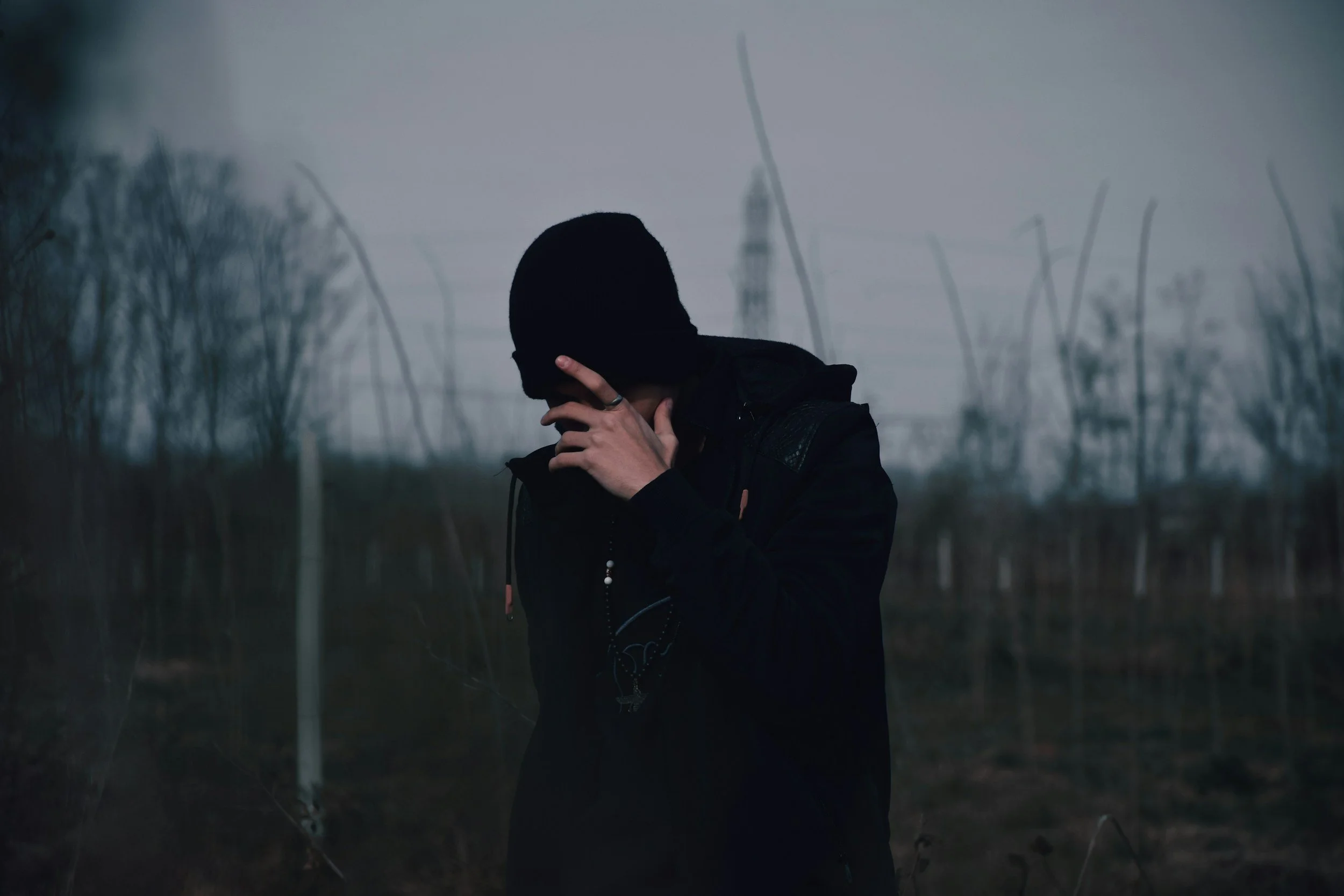 A person dressed in black with a hood covering their head, standing outdoors in a foggy, desolate area with leafless trees and power lines in the background, appearing contemplative or distressed.