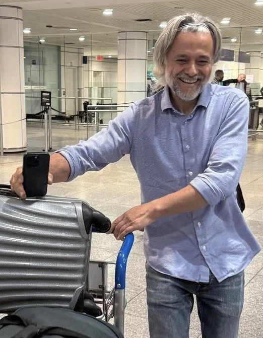 A smiling man with long gray hair and a beard, wearing a light blue long-sleeved shirt, is standing in an airport terminal. He is holding a mobile phone with one hand and pushing a luggage cart with the other.