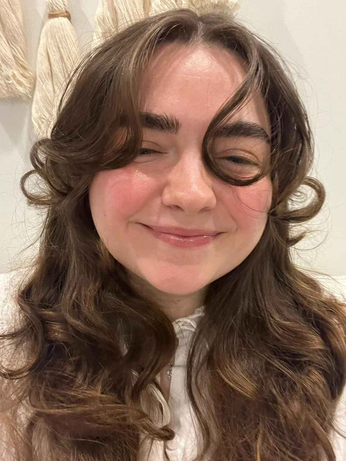 A smiling woman with long, curly brown hair, closed eyes, and light skin, wearing a white top, standing in front of a white wall and beige curtain.