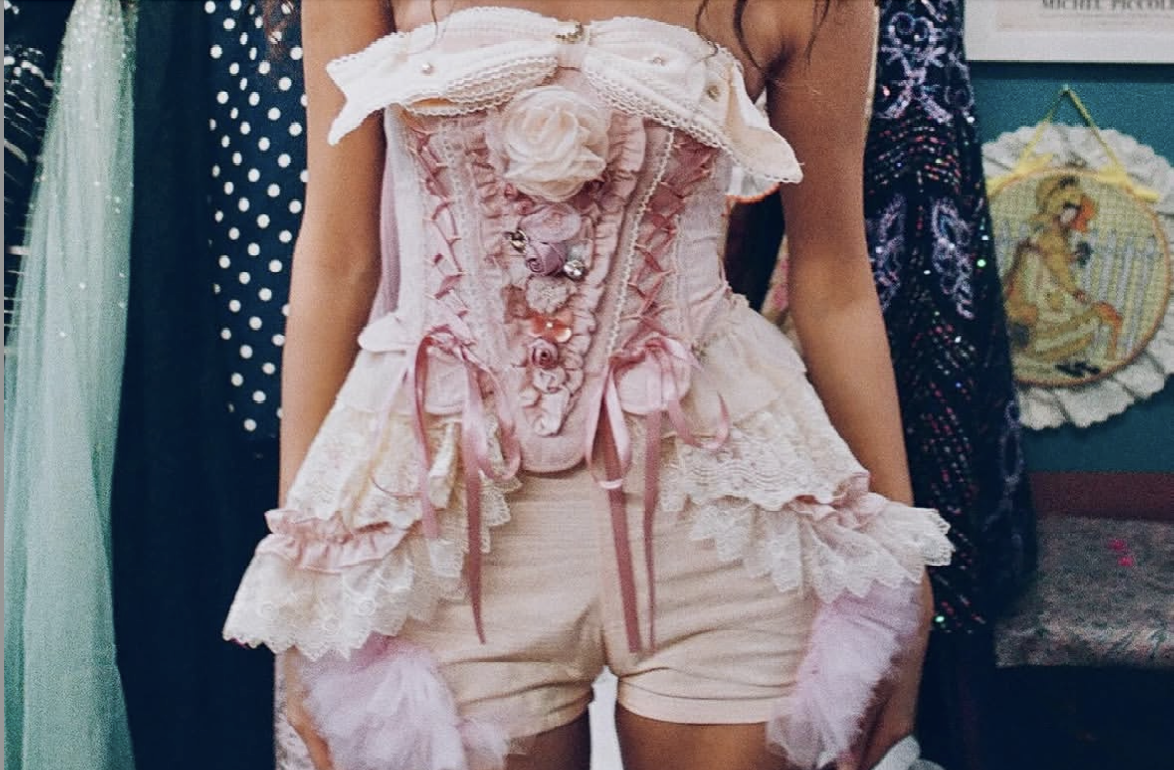Person wearing a pink and cream lace corset top with ruffles and bows, paired with matching shorts featuring lace, ruffles, and pink tulle accents, at a costume or vintage-themed event.