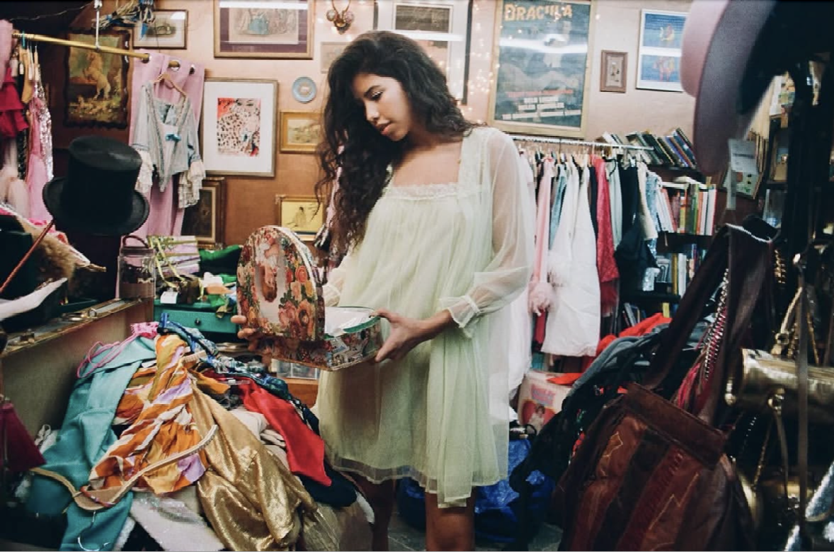 A woman shopping at a thrift store, holding a floral purse and examining clothes on a table filled with colorful garments.