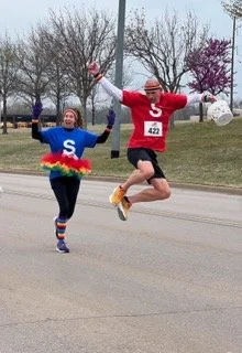 Two people dressed as superheroes with large letter 'S' on their chests, jumping and running on a street during daytime. One wears a blue costume with rainbow-colored fringe, and the other wears a red costume with the number 422. Trees and purple flowering trees are in the background.