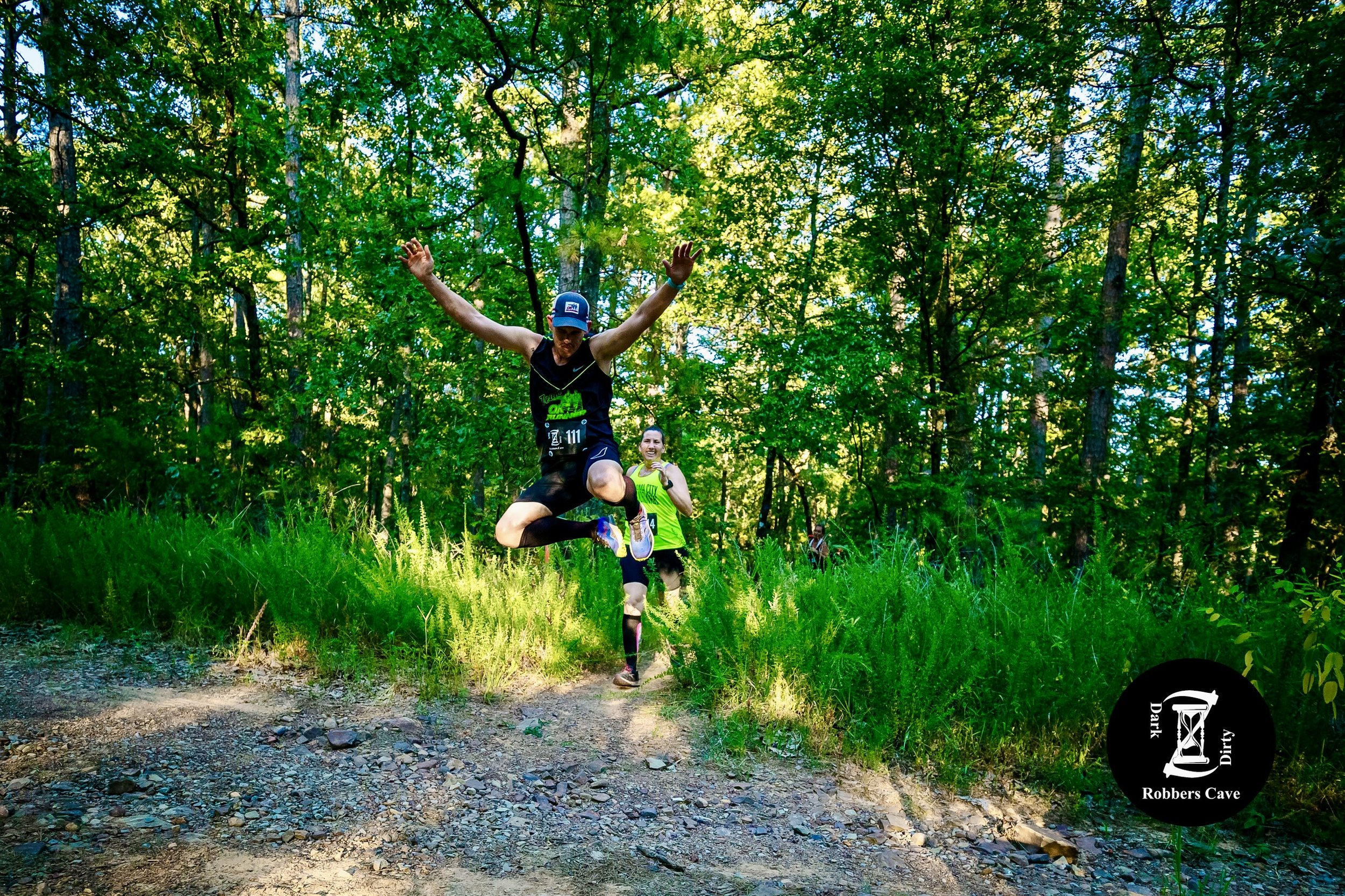 Two runners, a man and a woman, participating in an outdoor trail race through a green forest. The man in front is jumping as he runs, wearing a black tank top, black shorts, and a blue hat. The woman behind him is smiling, wearing a bright green top and black shorts.