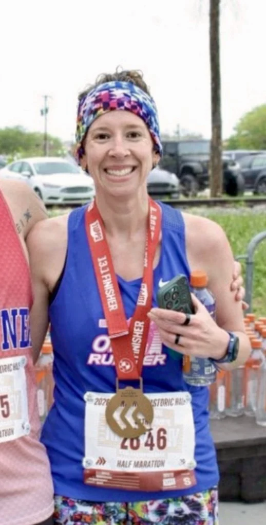 A smiling woman wearing a blue race shirt, colorful headband, and a finisher medal around her neck, holding a phone and a water bottle, after completing a half marathon.