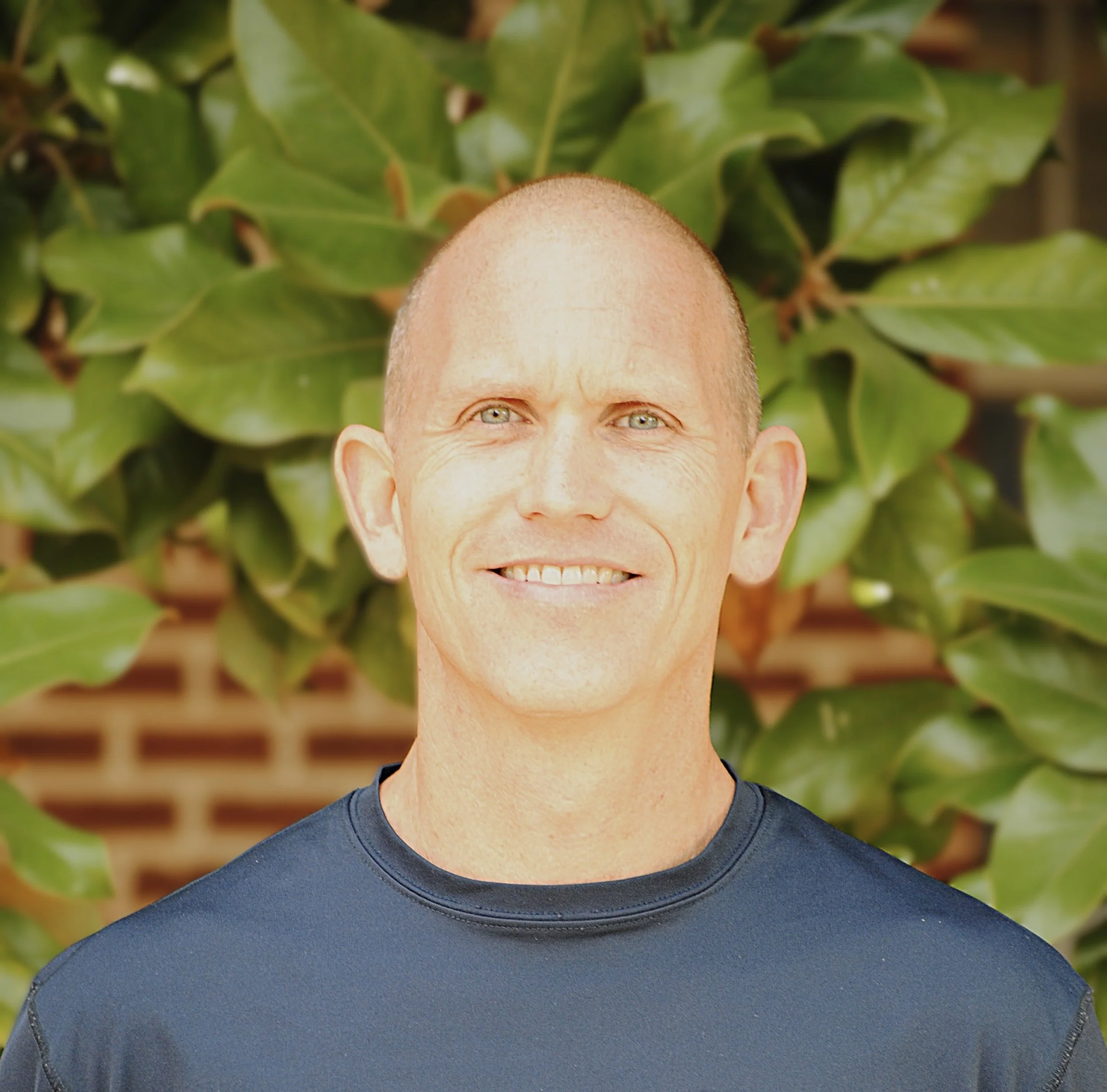Close-up of a smiling man with blue eyes, wearing a dark blue shirt, outdoors with green leaves in the background.