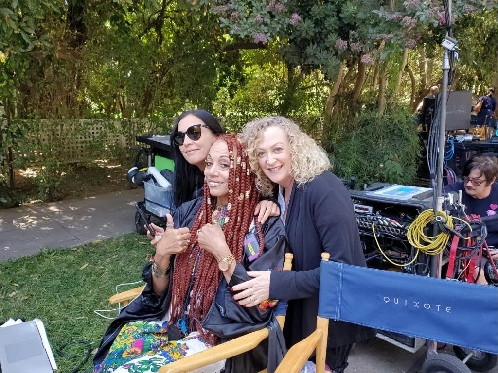 Three women on a film set outdoors, one with long red dreadlocks, sitting in a director's chair with a