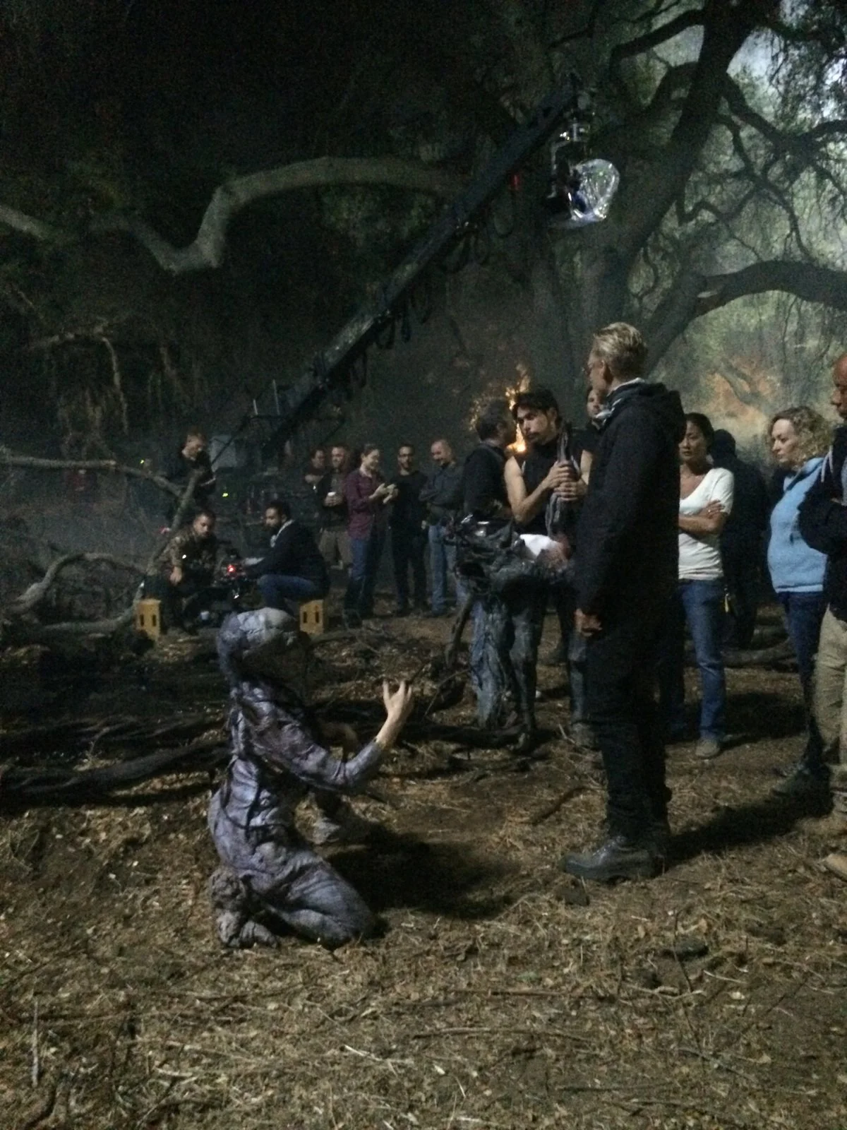 A group of people gathered outdoors at night, with some kneeling and others standing, around a fallen tree in a wooded area. The scene appears to be a film or photo shoot, with equipment and a large crane overhead, and a person in costume kneeling on the ground.