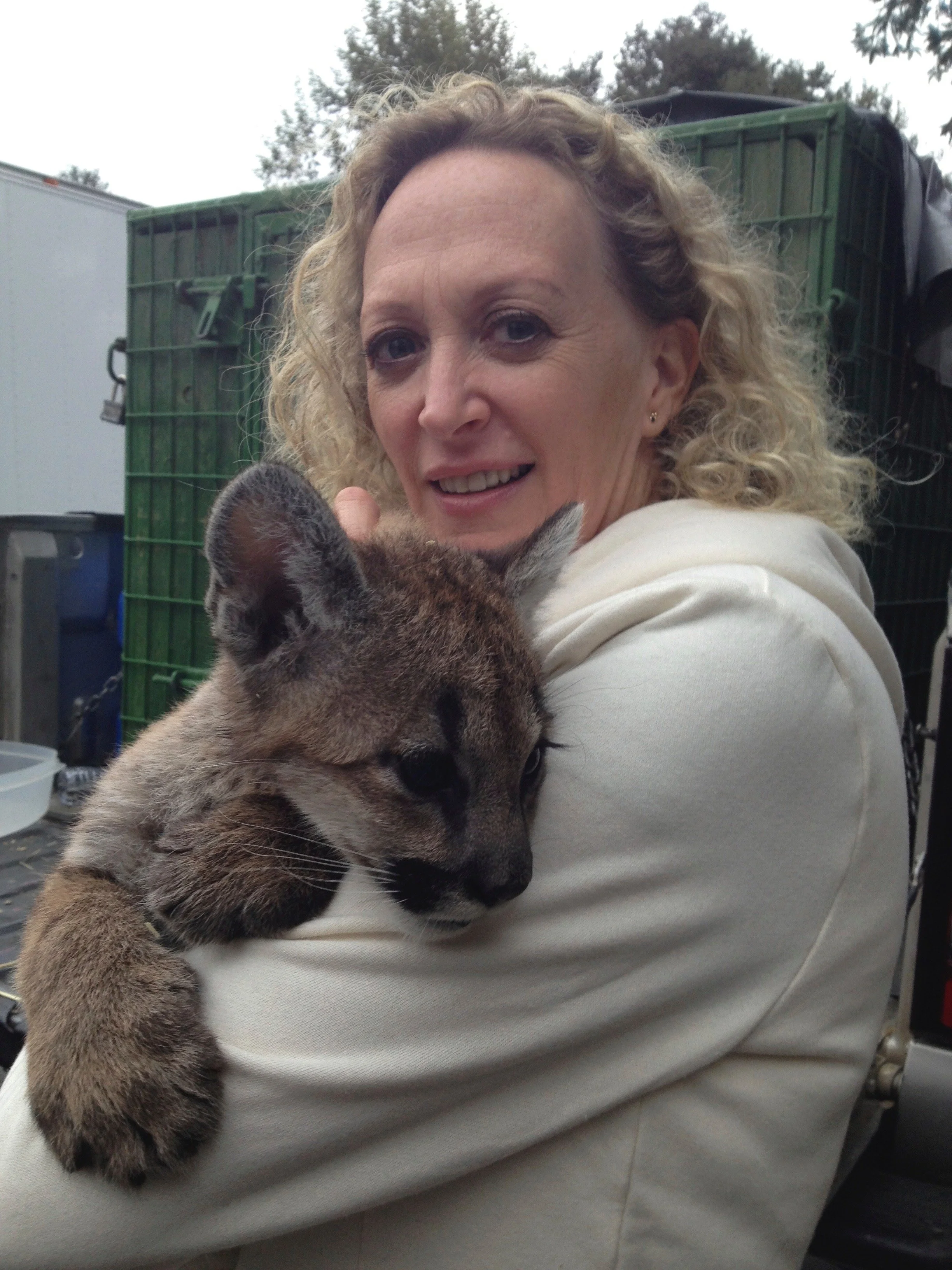A woman with curly blonde hair holding a tiger cub in her arms. The scene appears to be outdoors with trees and a green cage in the background.