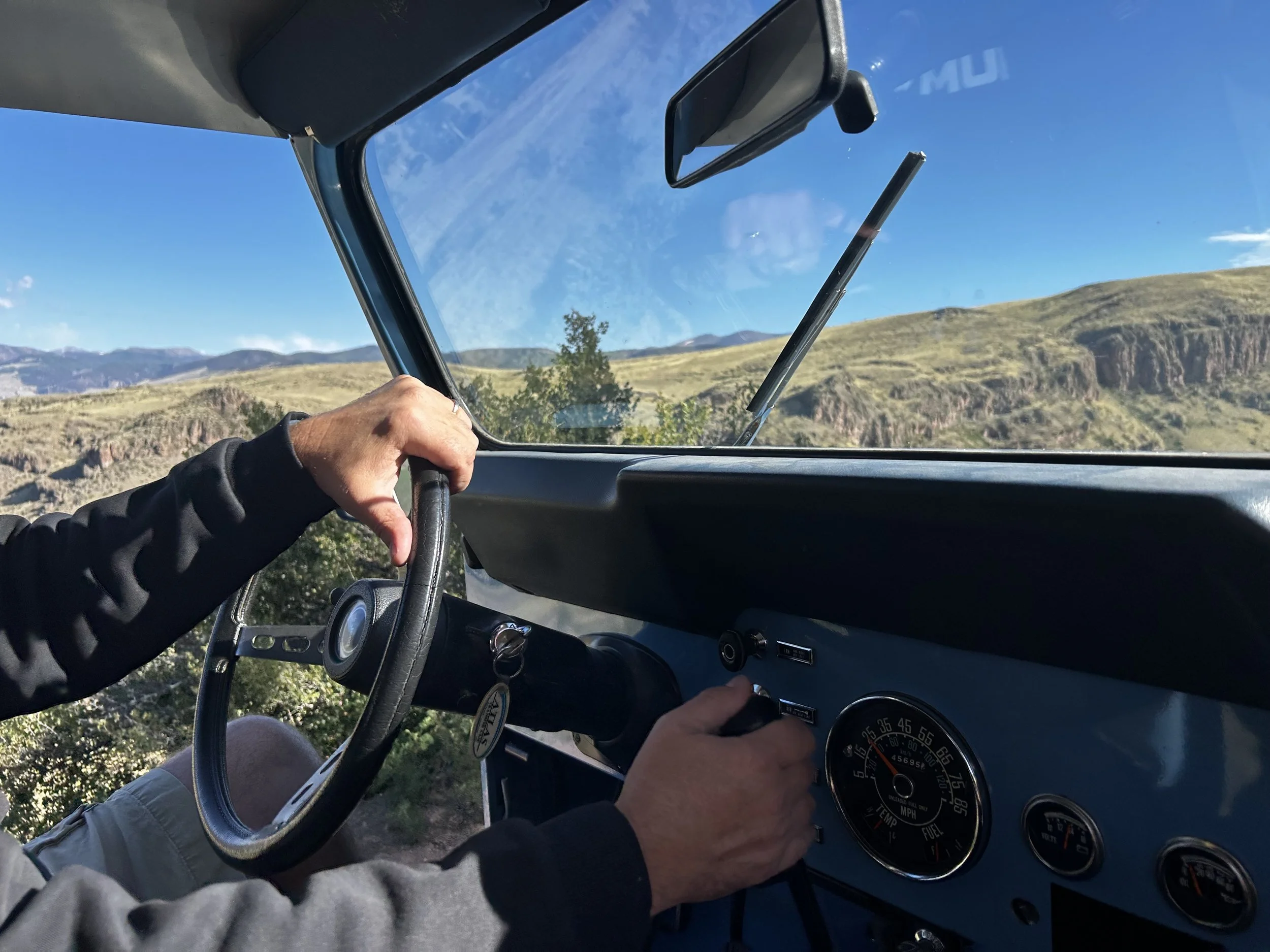 Inside a vehicle, a person's hand is adjusting the speedometer while another hand is on the steering wheel. The dashboard shows the speed in miles per hour, and the view outside through the windshield reveals a scenic landscape with hills, trees, and a clear blue sky.