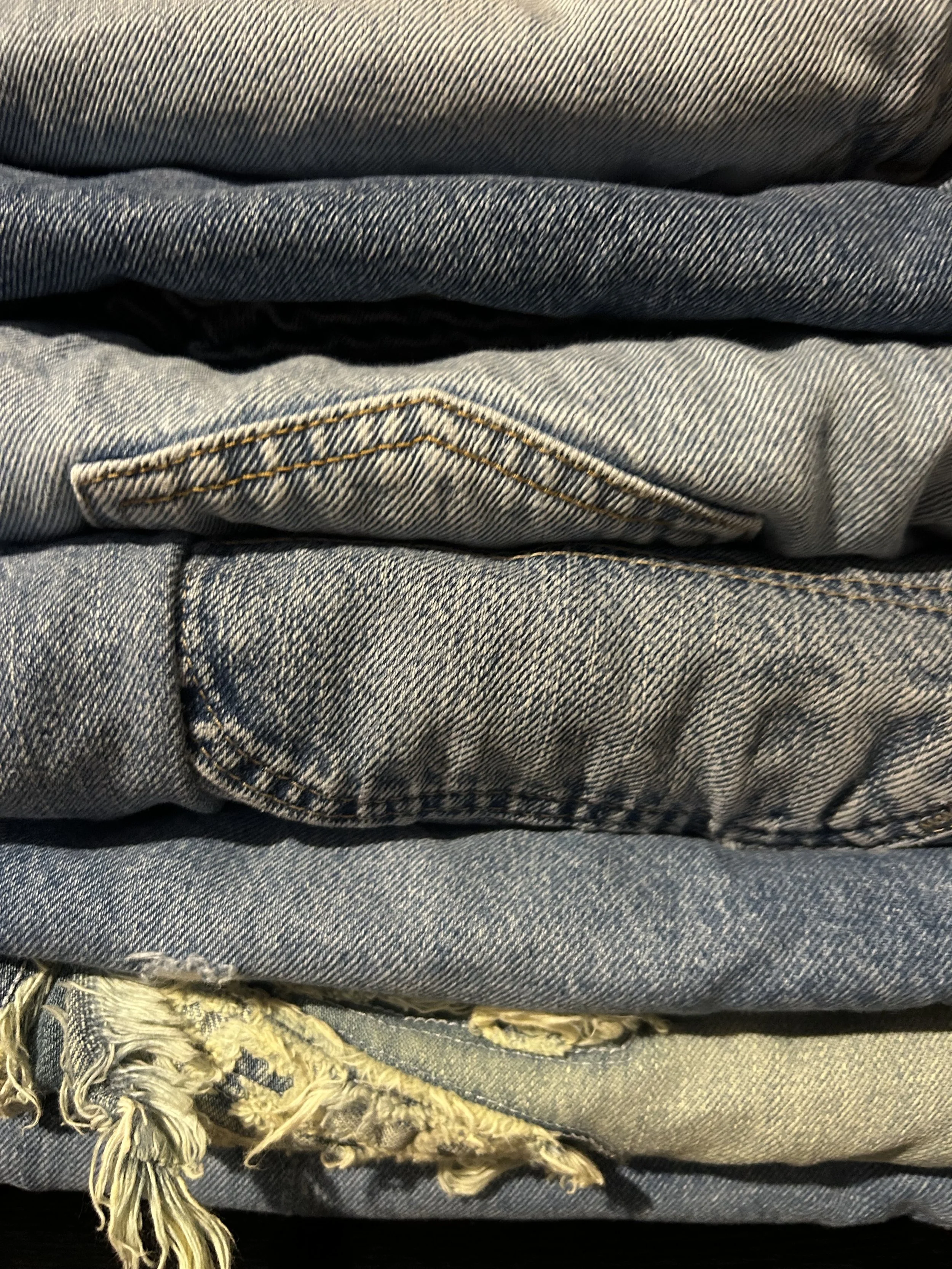 Stacked denim jeans in various shades and styles, including light, medium, and dark washes, some with frayed hems, arranged in layers.