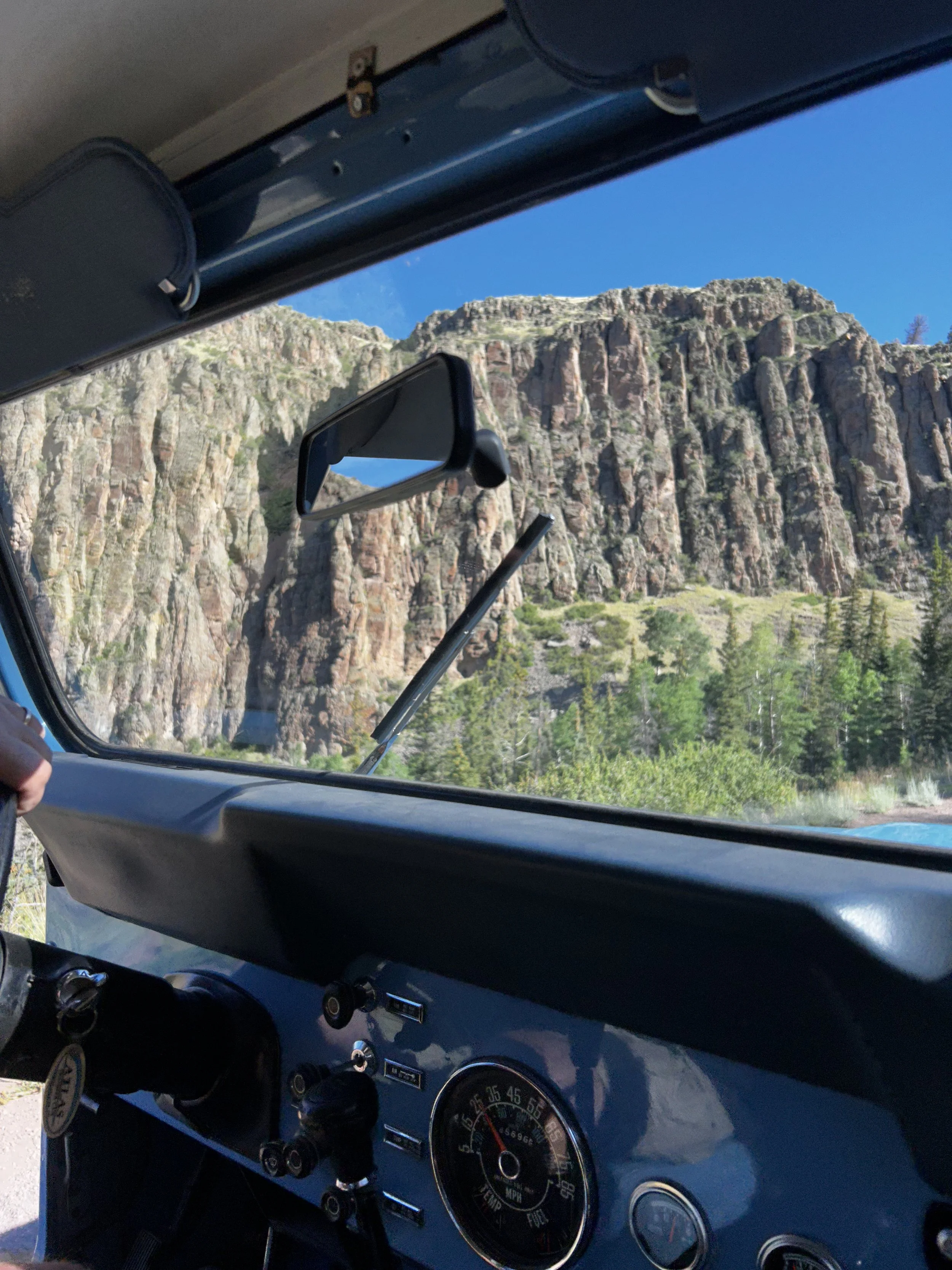 Inside a vehicle, looking out the windshield at a mountainous landscape with a clear blue sky, rocky cliffs, and green trees.