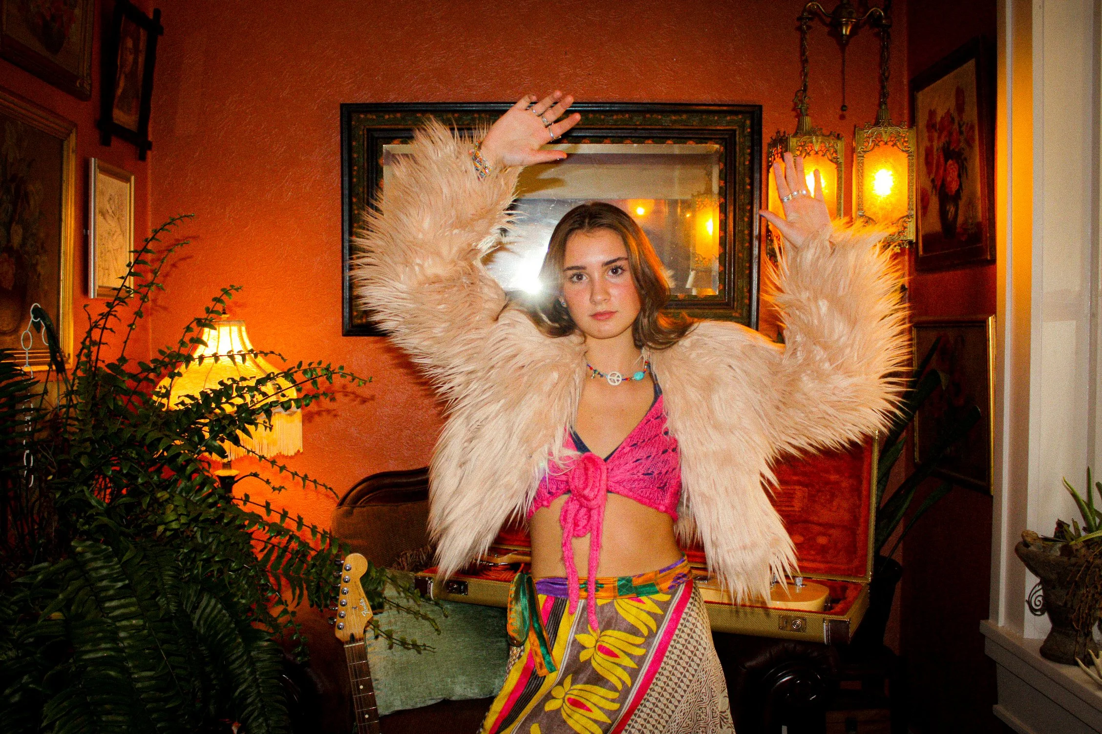 Young woman wearing a fluffy white jacket with raised arms in a warmly decorated room, surrounded by plants and artwork, with a guitar in the background.
