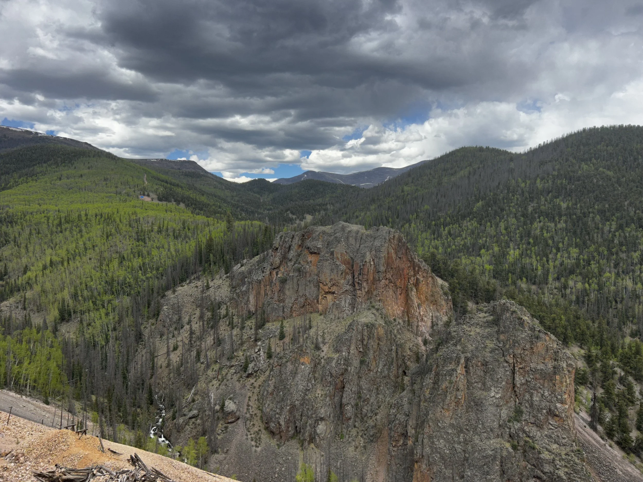 A mountain landscape with a cloudy sky, lush green trees on the hills, rugged rocky formations, and a small river at the bottom.
