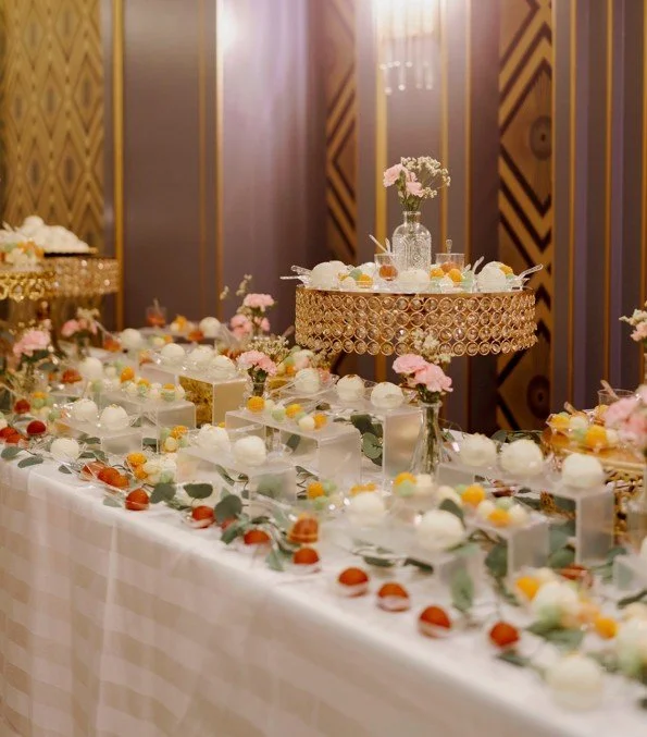 Decorative table with pink flowers, white desserts, and colorful small treats, set against a softly lit, patterned wall.