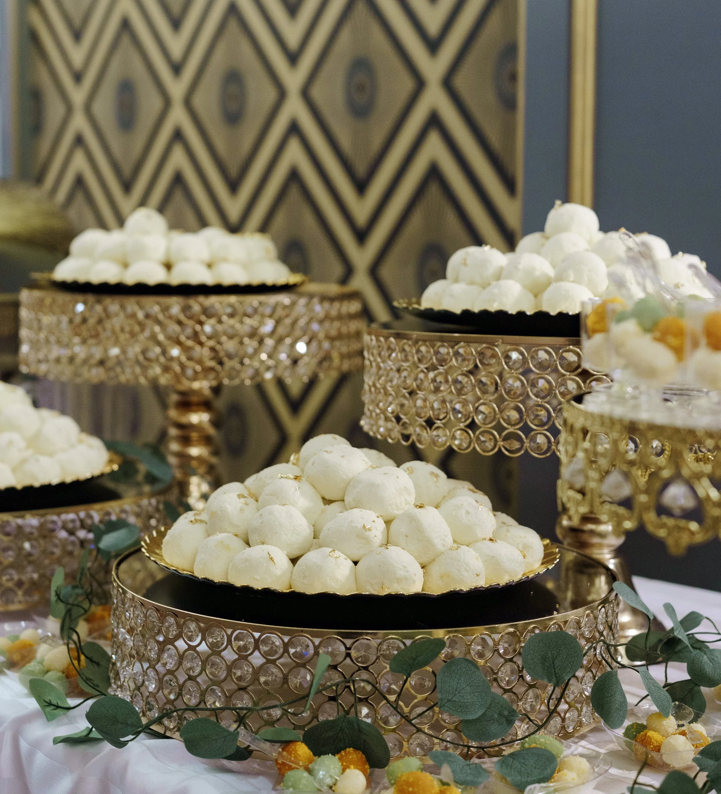Two round white desserts on a decorative golden tray, central to an elegant dessert display, with a patterned wallpaper background.