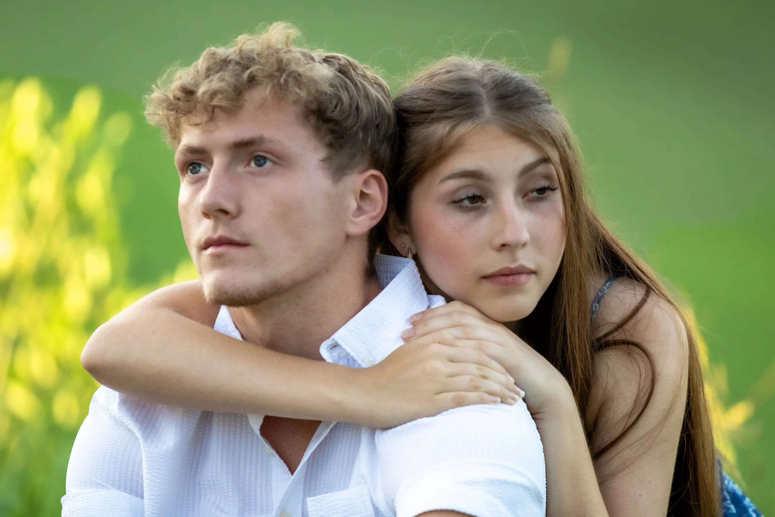A young man with curly blonde hair and a woman with long brown hair are close together outdoors, with the woman gently resting her arms over the man's shoulders. They have serene expressions and are surrounded by a blurred green background of nature.