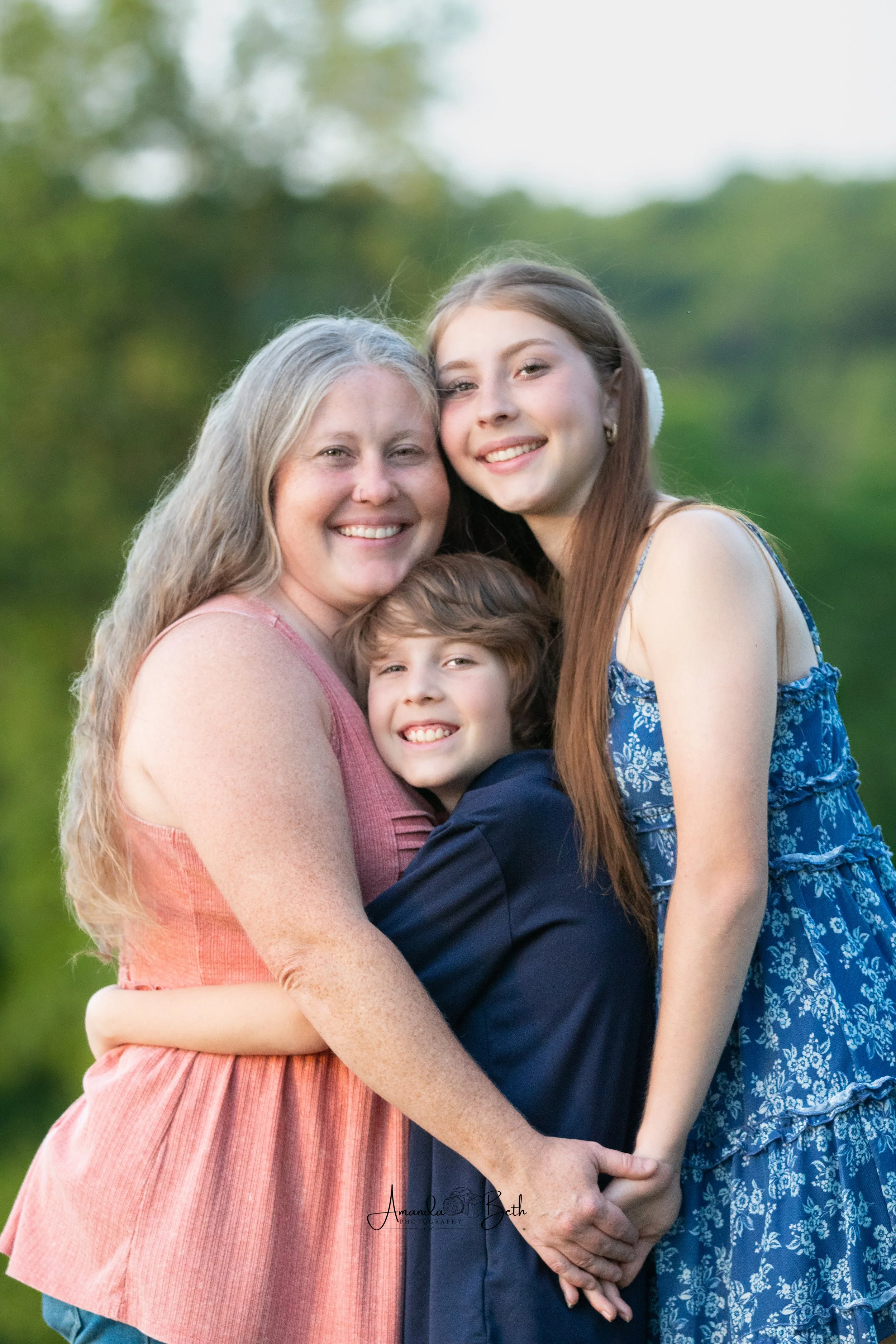 A mother and two children hugging outdoors with blurred green trees in the background, smiling at the camera.