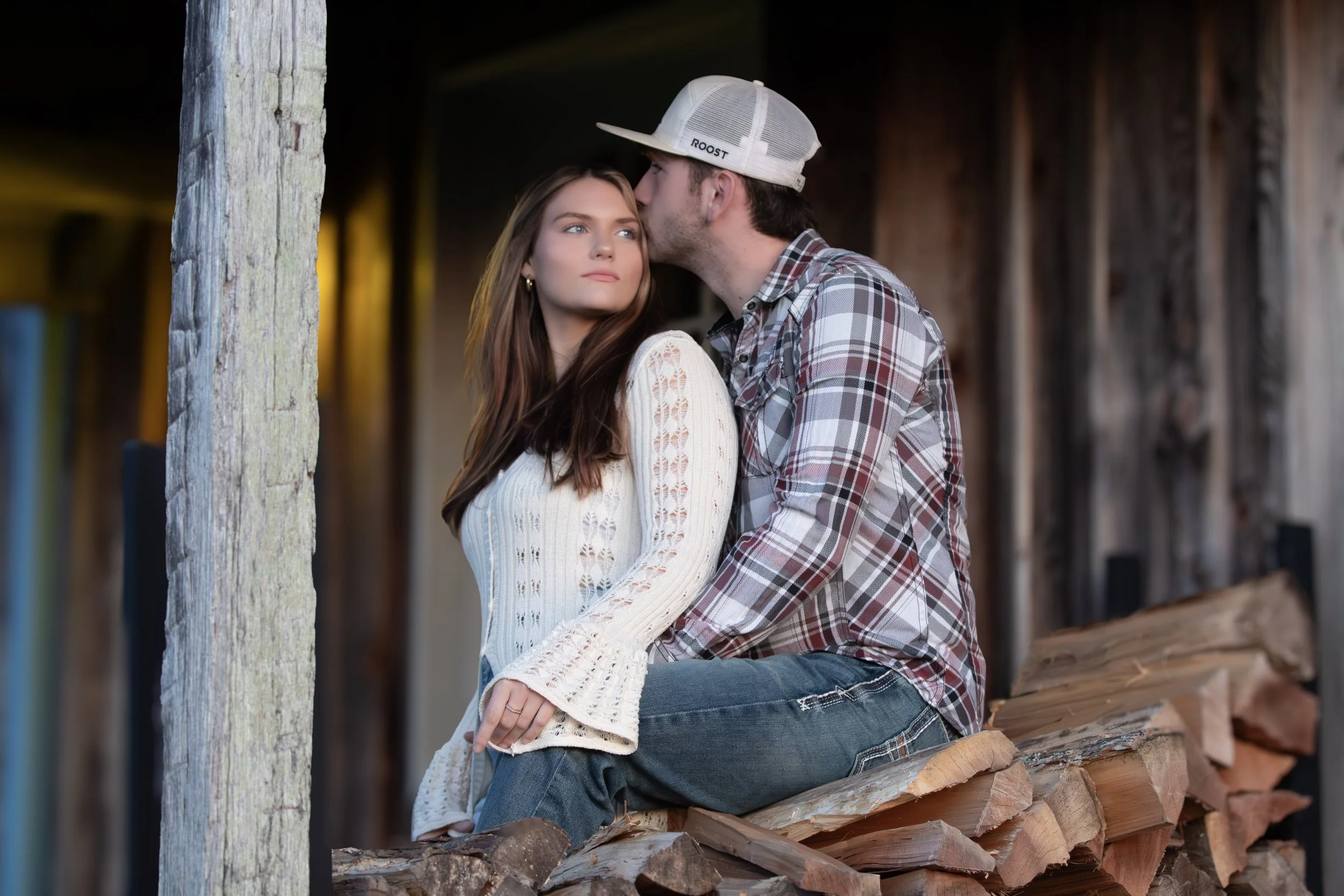 A young couple sitting on chopped firewood, with the man kissing the woman's forehead. The woman has long, brown hair and is wearing a white knitted sweater. The man is wearing a plaid shirt, jeans, and a baseball cap. They are outdoors with a wooden structure in the background.