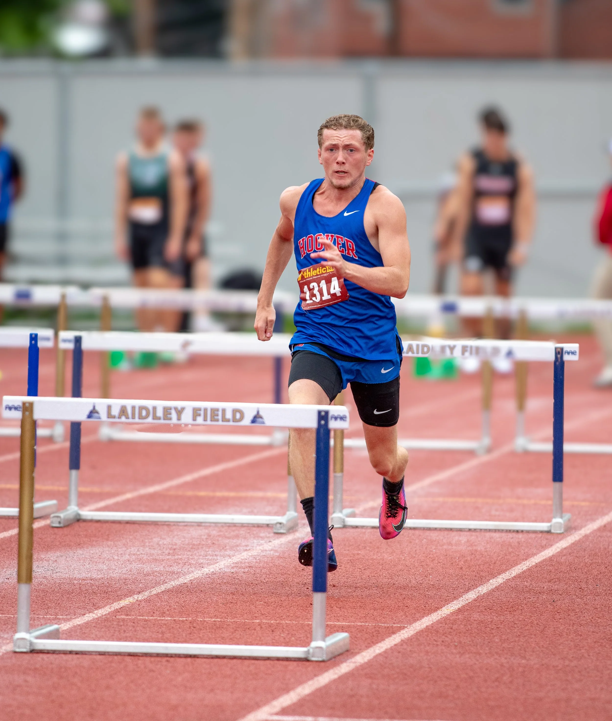 Male athlete in a blue athletic shirt and black shorts running and hurdling on a red track with hurdles labeled "Laidley Field" in an outdoor stadium. Several other runners are blurred in the background.