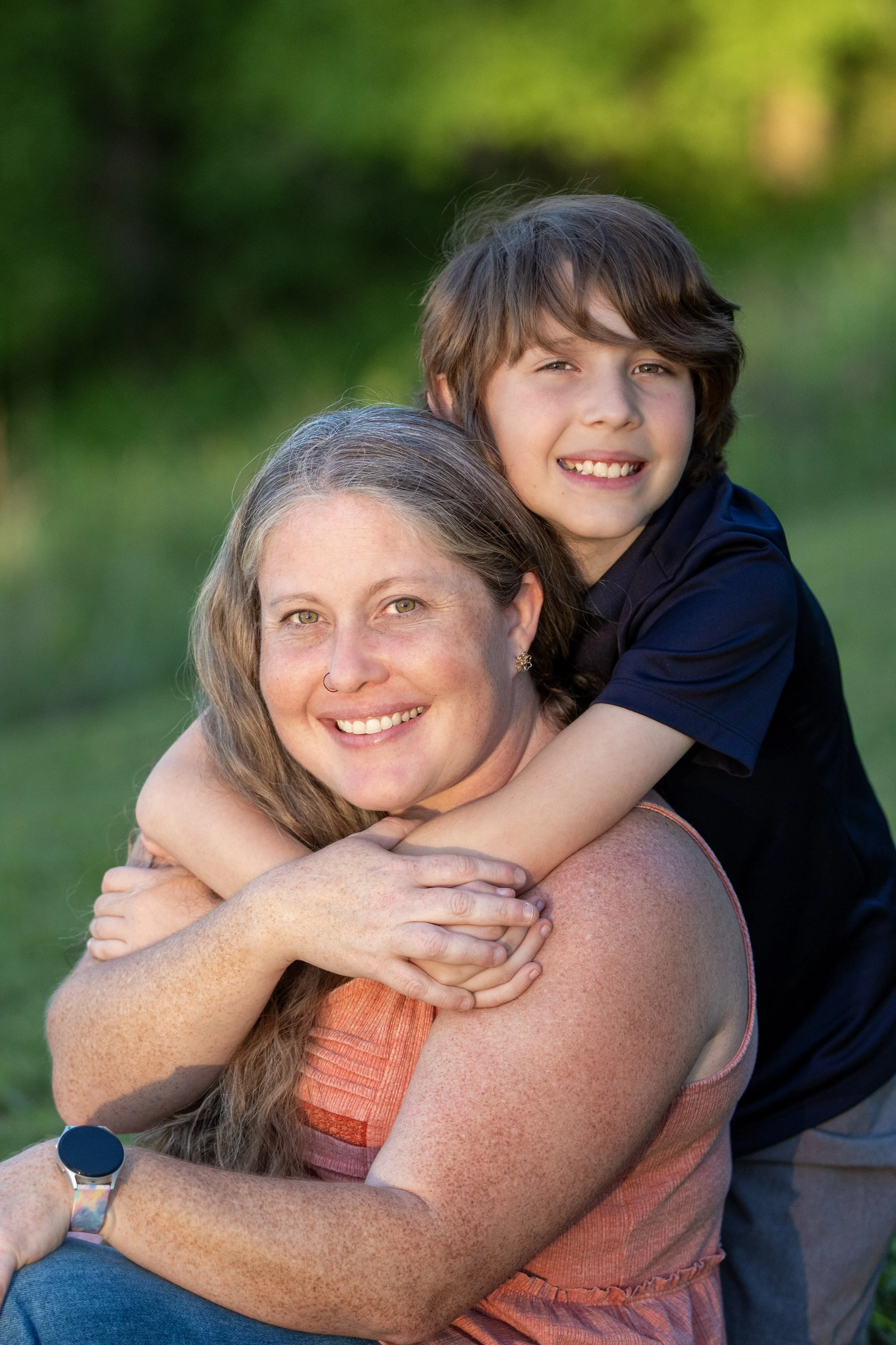 A woman with long hair and freckles smiling, and a boy with short brown hair and a big smile, hugging each other outdoors with trees in the background.
