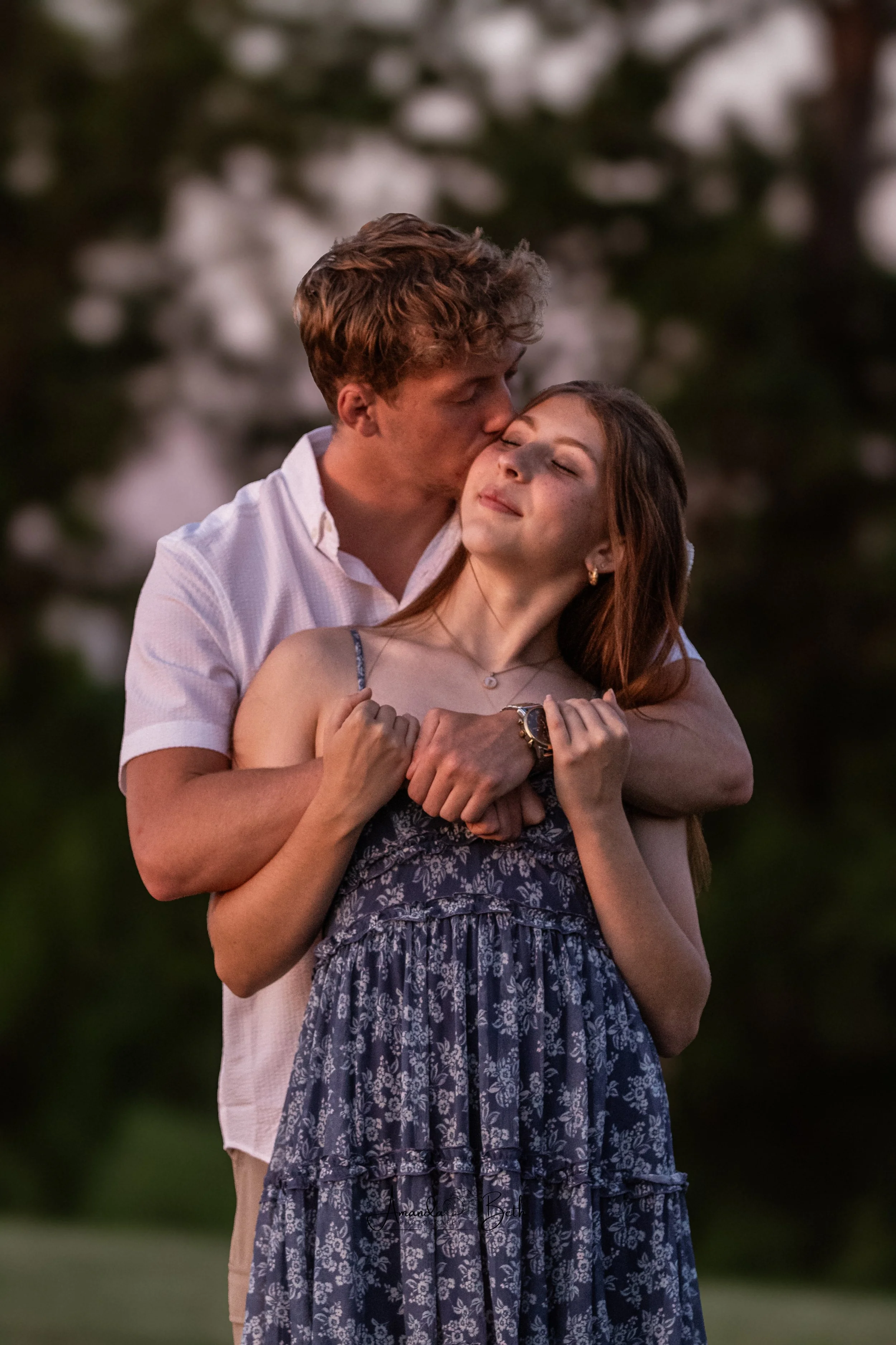A young man with curly brown hair is kissing a young woman with brown hair and a floral dress on the forehead outdoors during sunset. The woman has her eyes closed and a serene expression.
