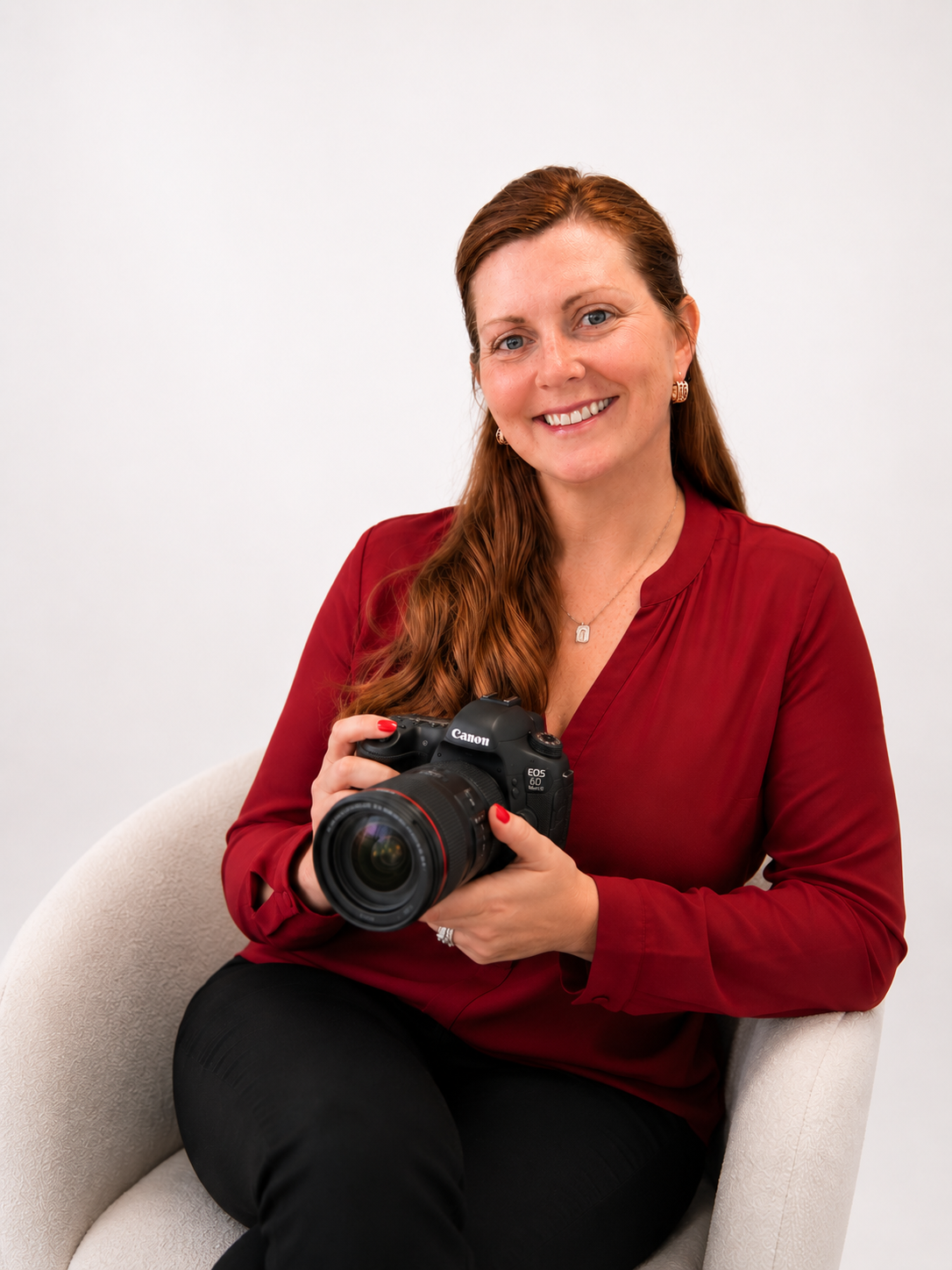 A smiling woman with red hair, wearing a red blouse and black pants, holding a Canon camera while sitting on a white chair.