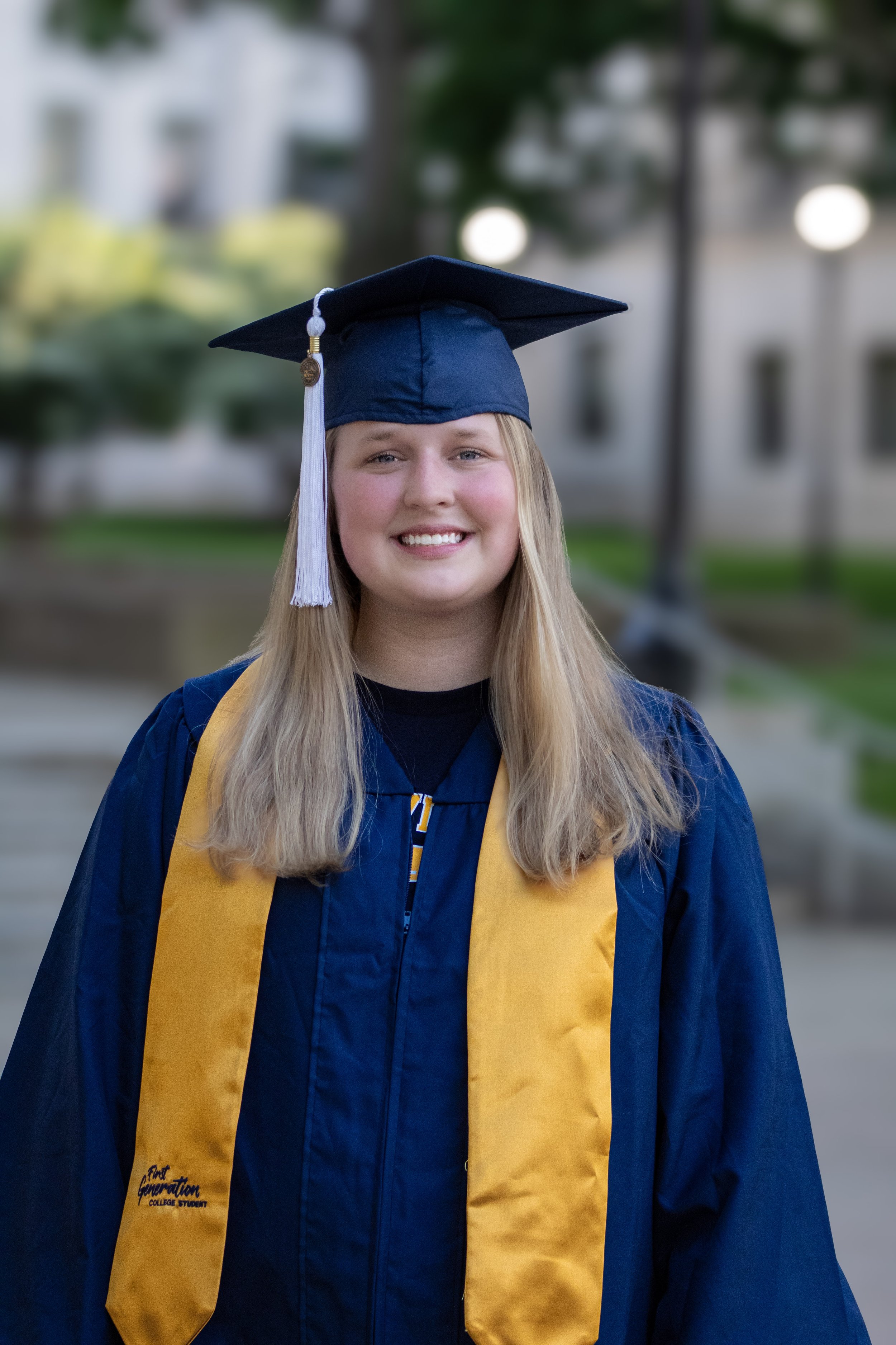 A young woman in a graduation cap and gown, smiling outdoors in front of a blurred background of trees and buildings.