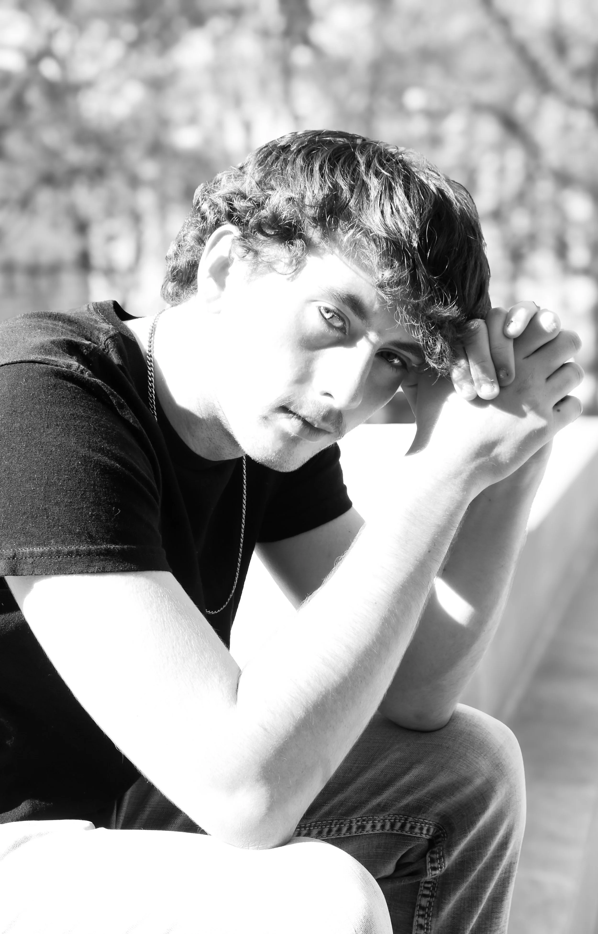 A black and white photo of a young man with short, curly hair, looking directly at the camera with a serious expression, resting his head on clasped hands in an outdoor setting.