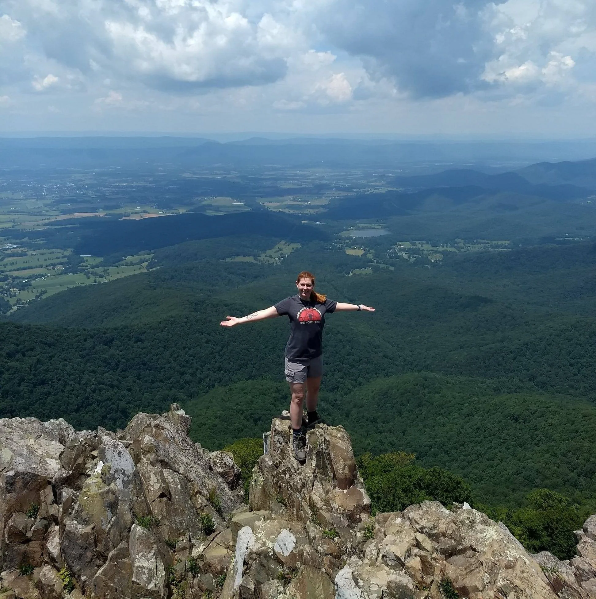 Woman standing on rocky ledge with arms outstretched atop a mountain, overlooking rolling hills, forests, and a valley under partly cloudy sky.