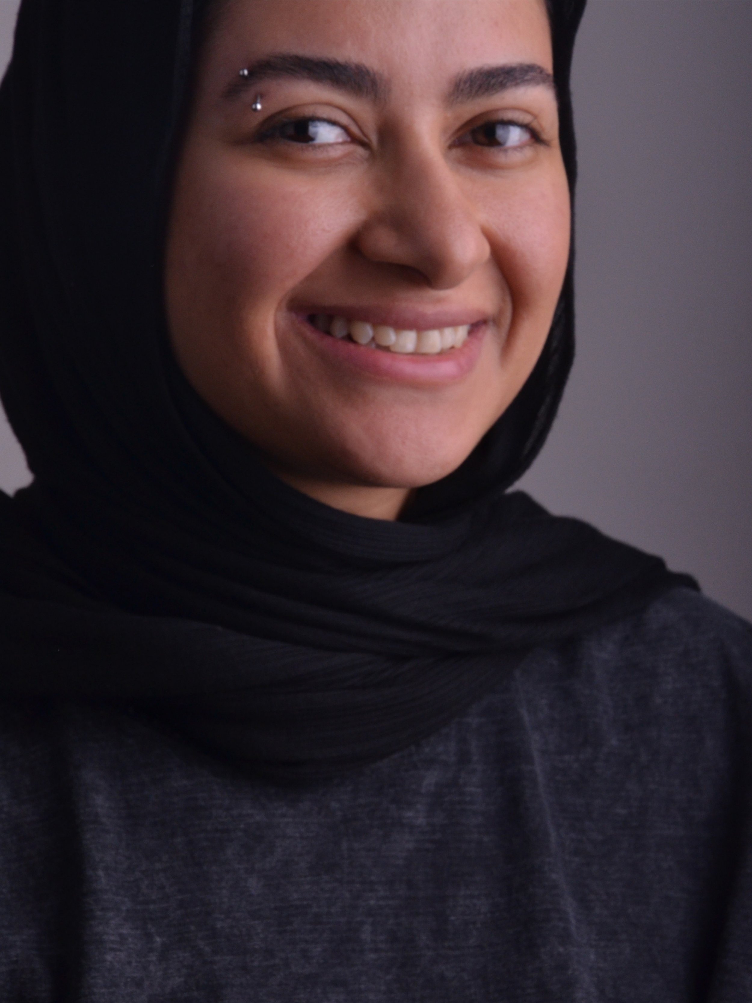 Close-up of a smiling woman with dark hair wearing a black headscarf and a dark sweater.