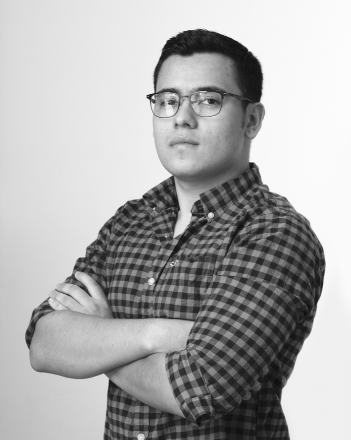 A young man wearing glasses and a checkered shirt with arms crossed, posing against a plain white background.