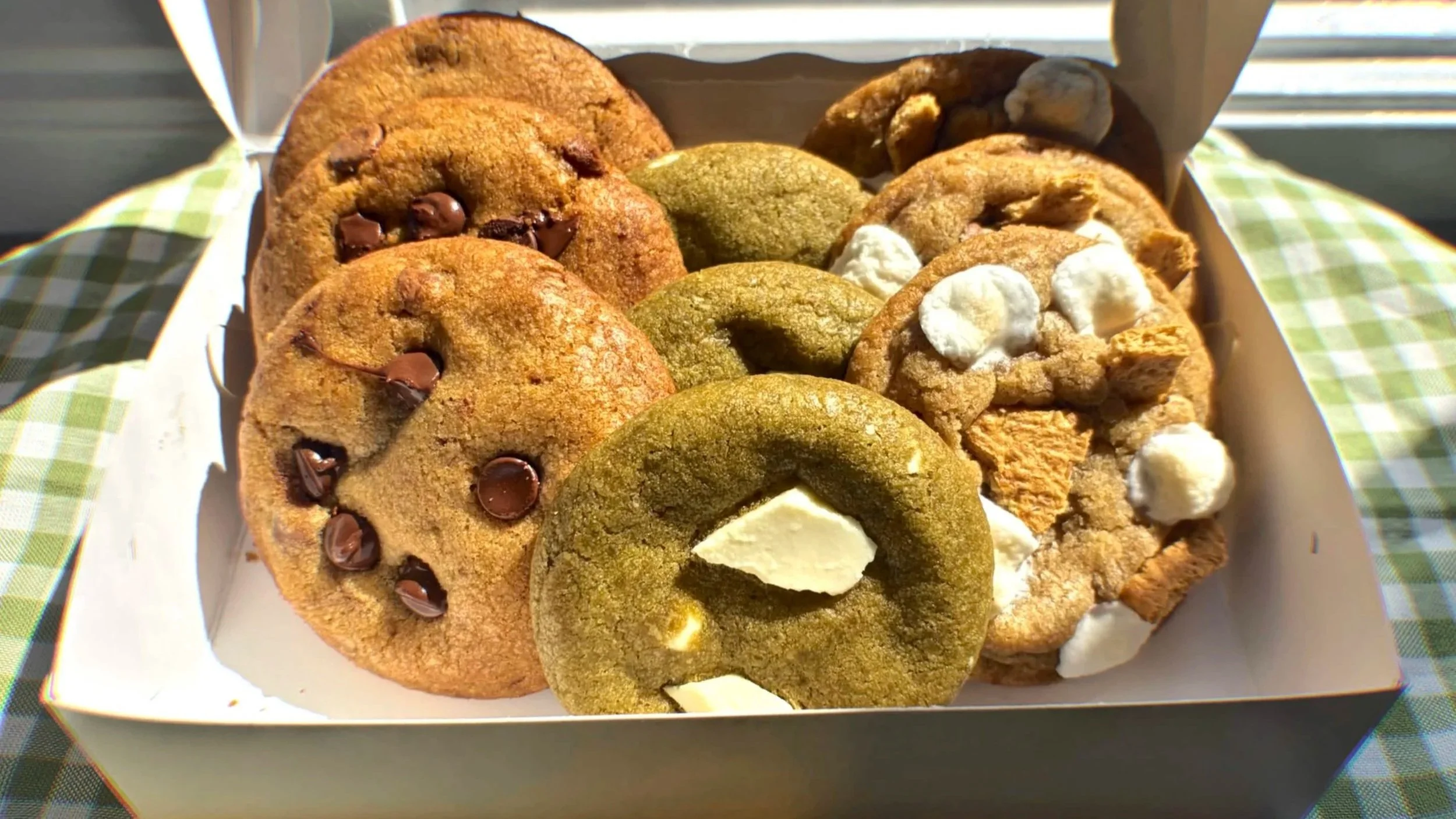 A white tray filled with a variety of cookies, including chocolate chip, green matcha, and cookies with white frosting, on a green checkered tablecloth.