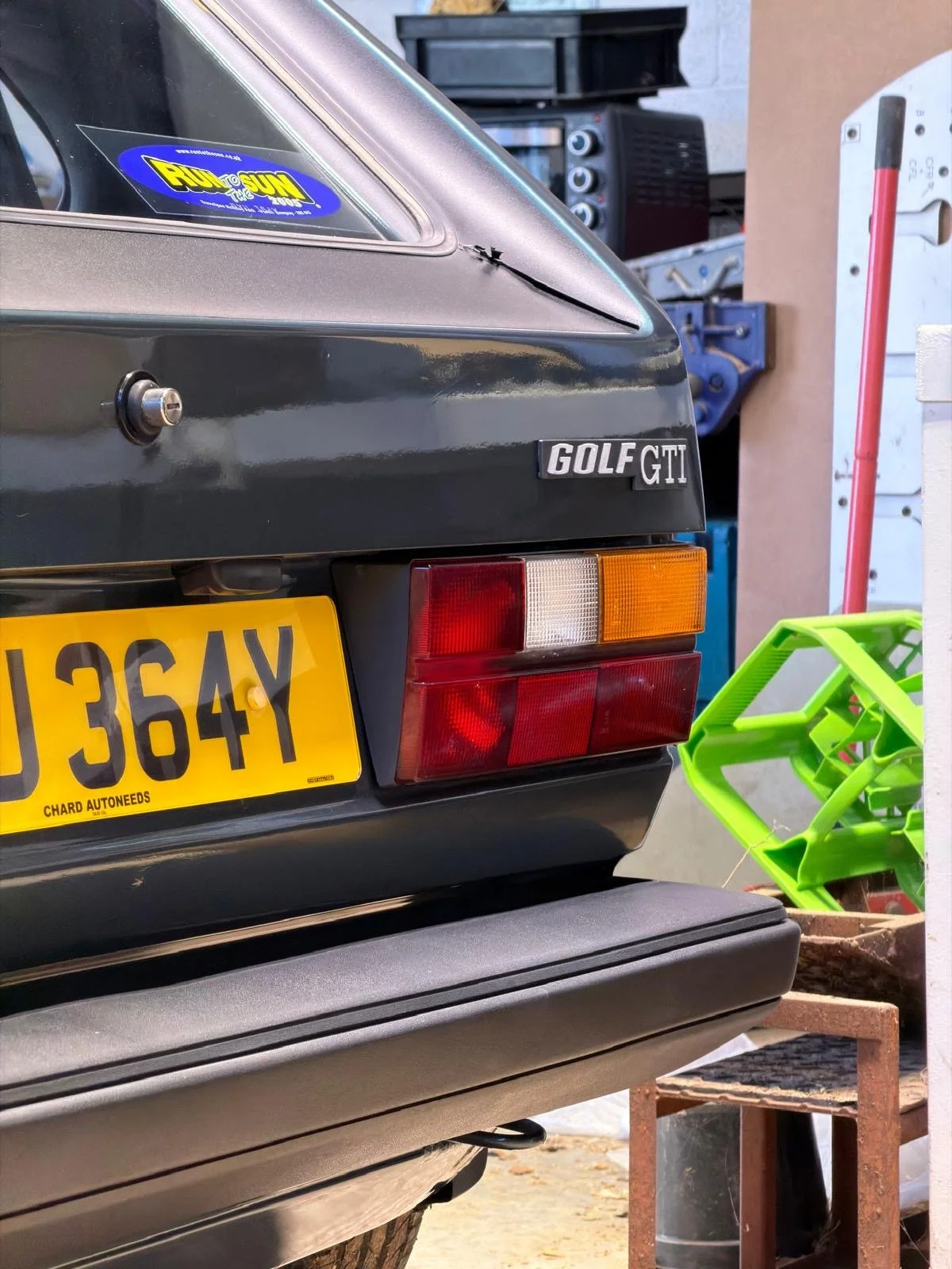 Rear of a gray vintage Volkswagen Golf GTI parked in a garage, showing license plate, tail lights, and badge, with various tools and objects nearby.