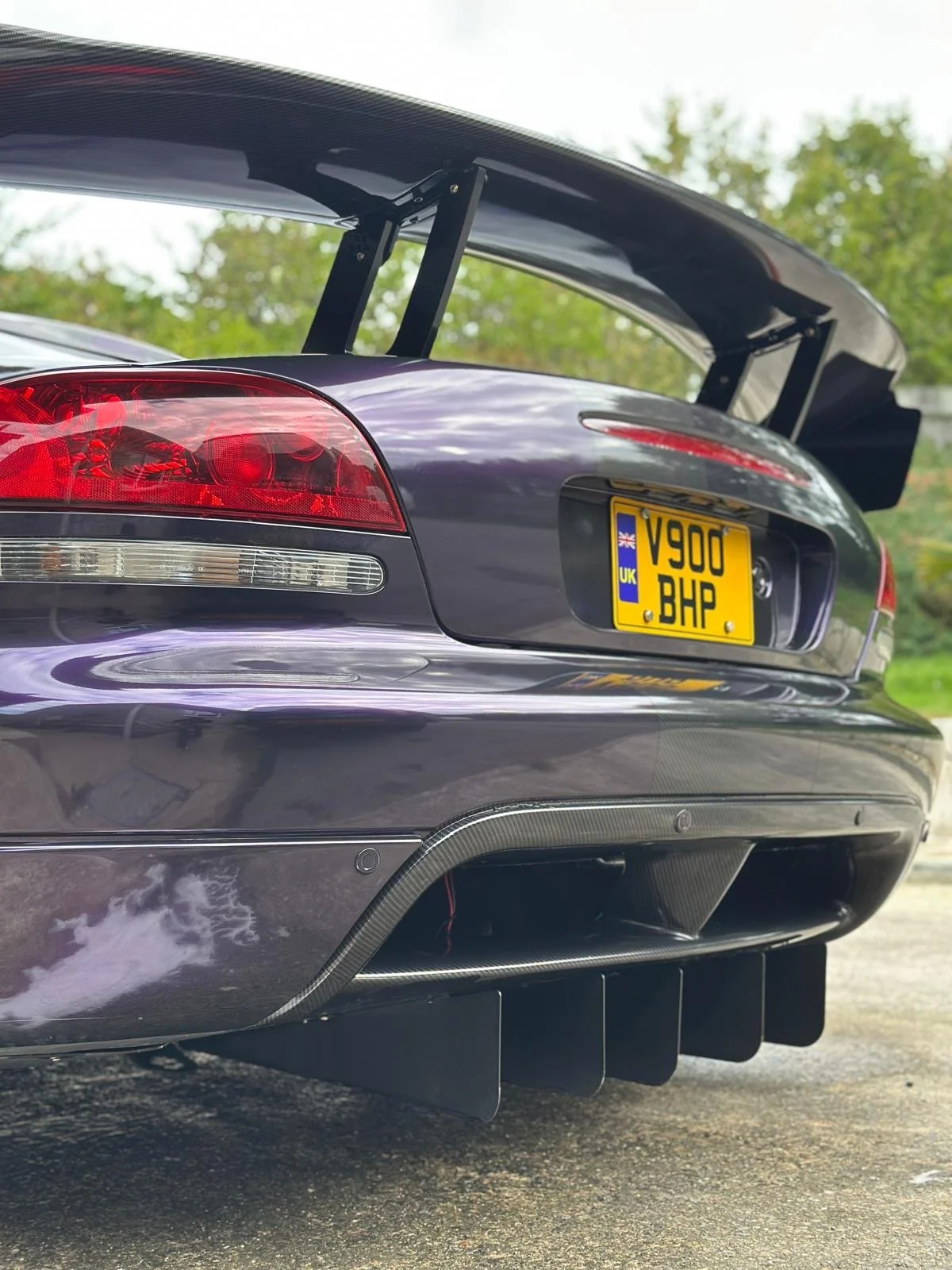 Close-up of the rear end of a black sports car with a large wing spoiler, custom carbon fiber rear diffuser, and custom license plate reading 'V900 BHP'.