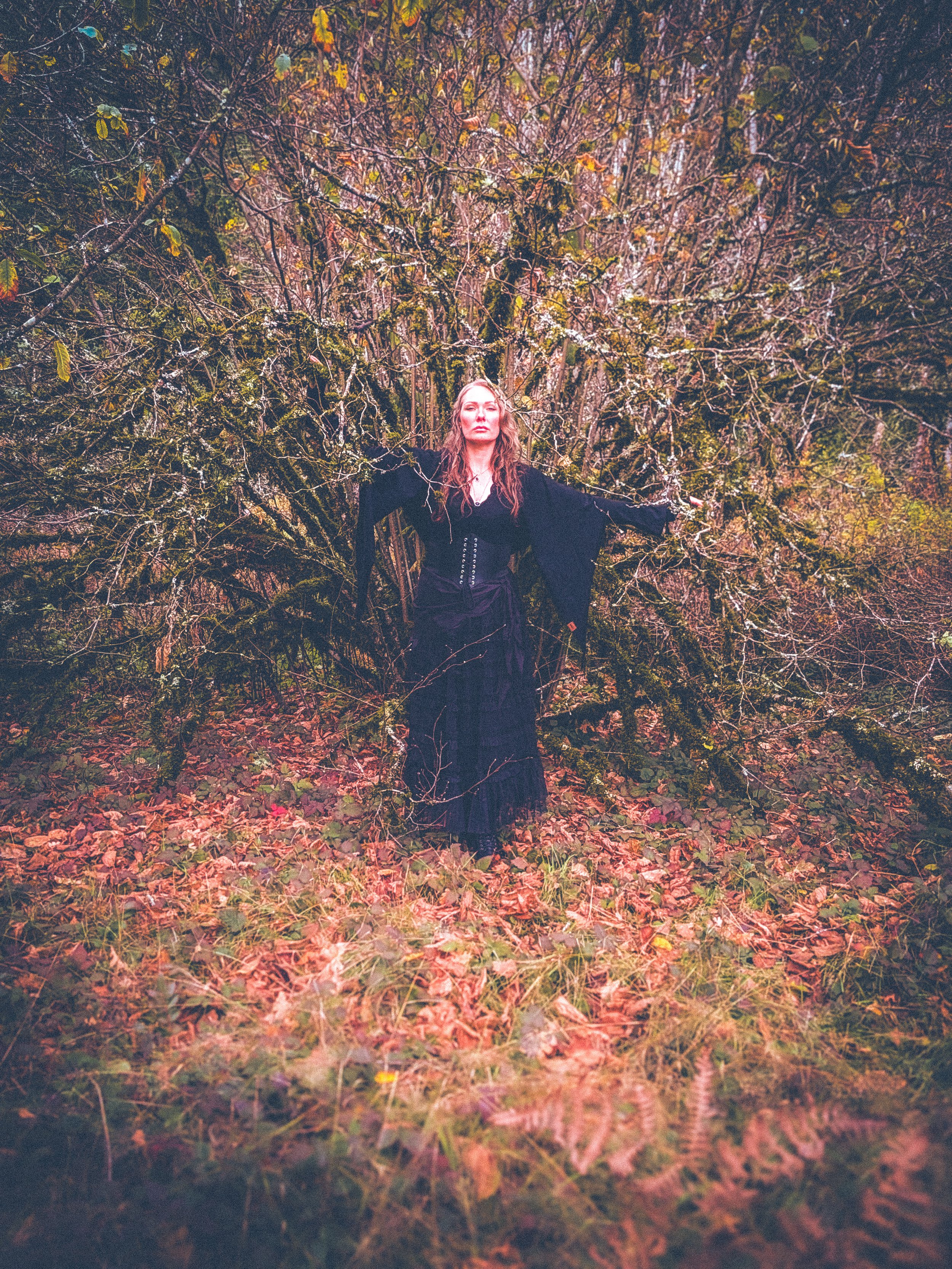 A woman with long, wavy hair, dressed in black, standing outdoors among bare, moss-covered trees and fallen autumn leaves.