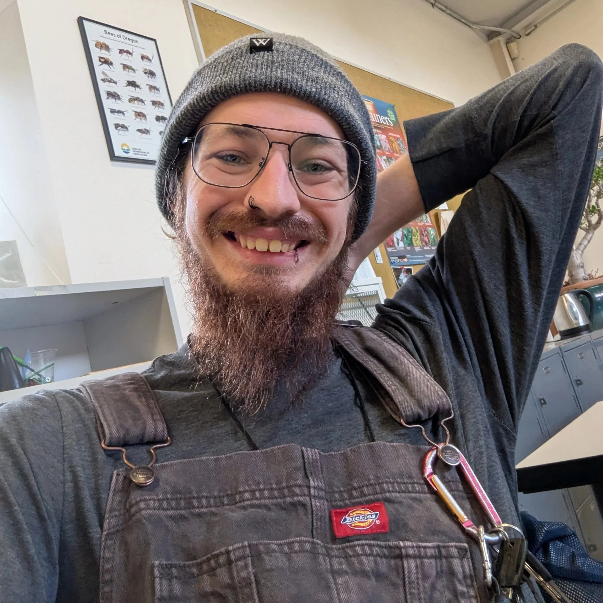 Close-up of a man with glasses, a gray beanie, and a beard, smiling, sitting in a room with posters and a white cabinet in the background.