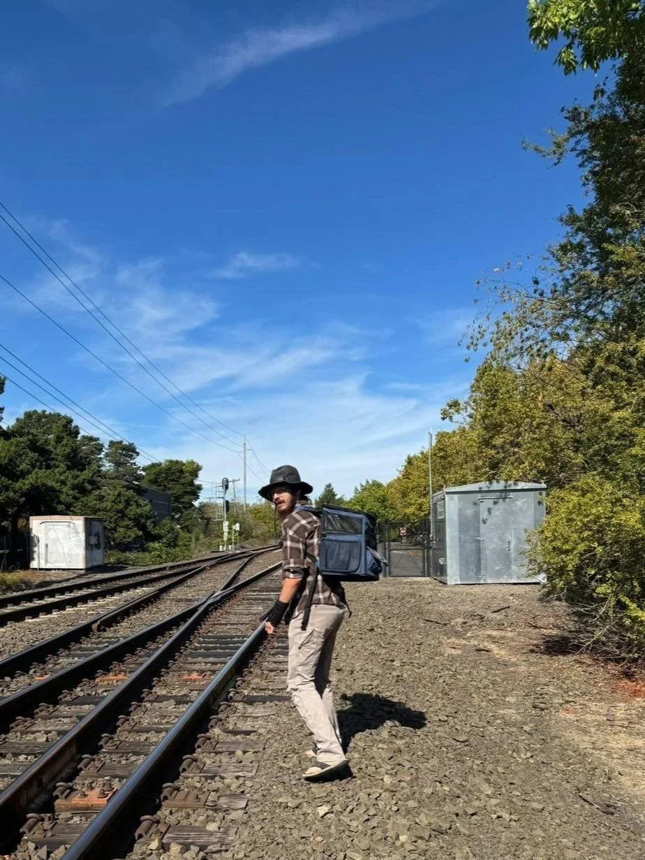 A person in a plaid shirt, beige pants, black hat, and sunglasses, carrying a backpack, walking along a railway track on a sunny day with trees and small structures nearby.