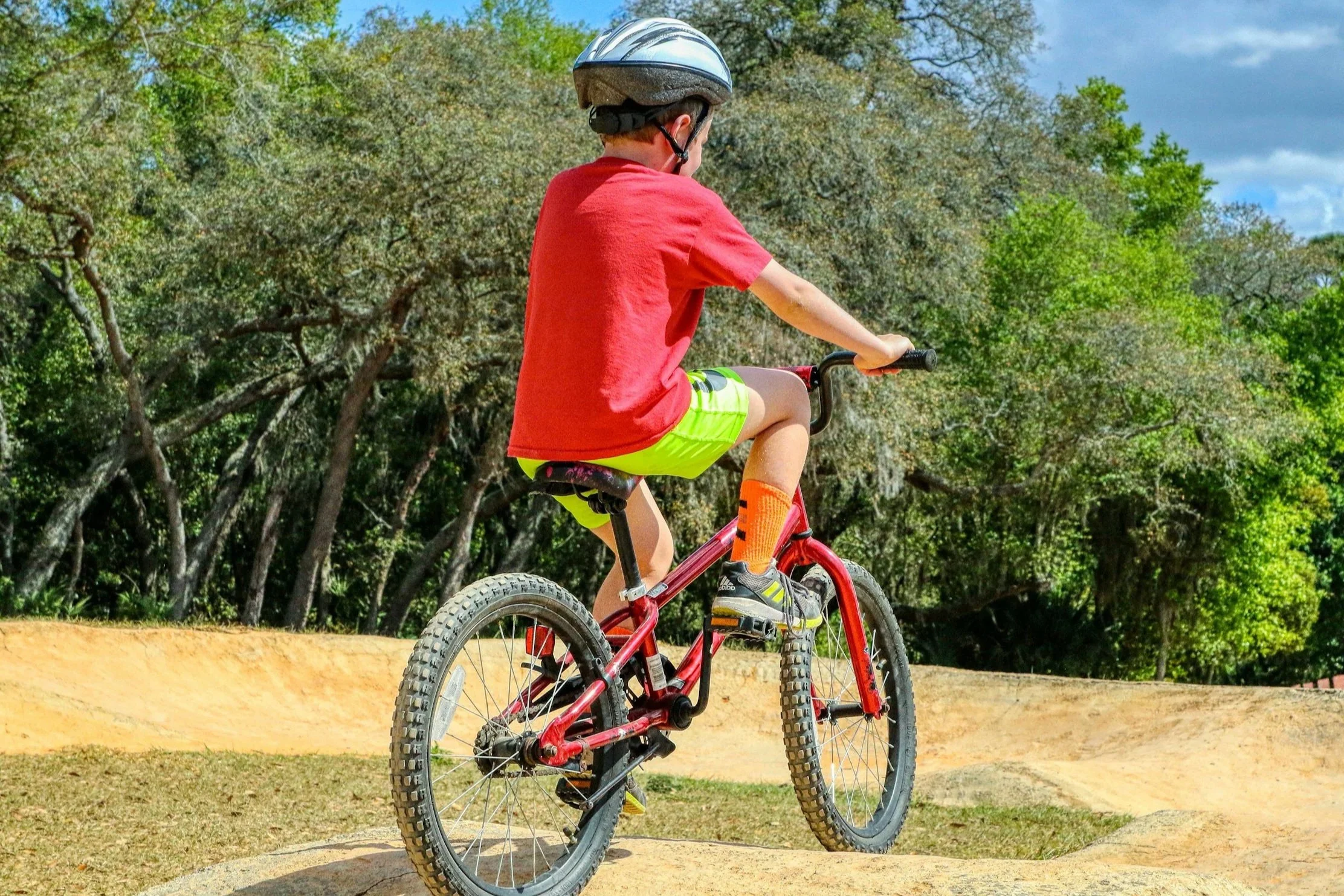 A young boy riding a red bicycle on a dirt path in a park with trees and a partly cloudy sky.
