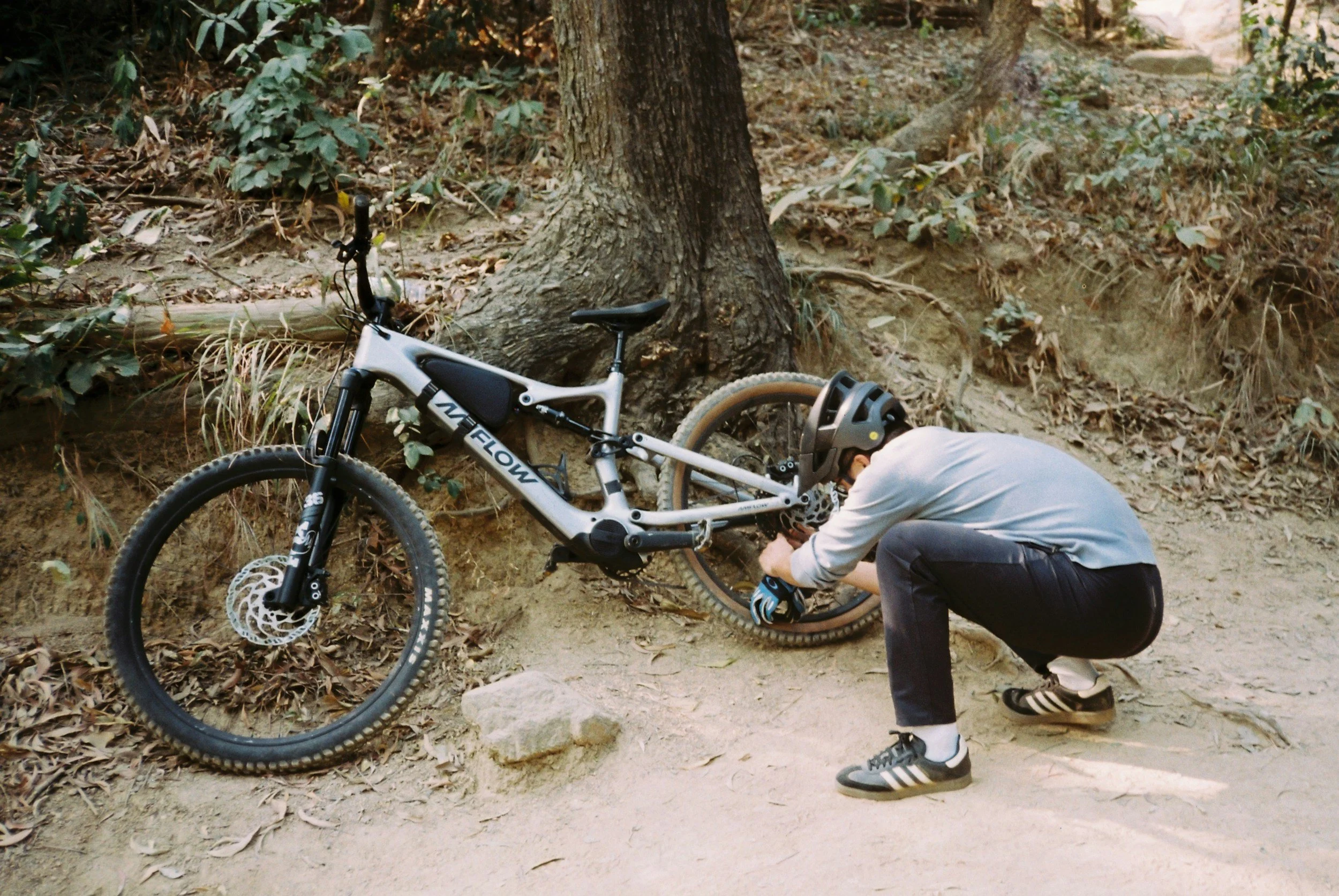 A person in a gray hoodie and black pants fixing or inspecting a white mountain bike next to a tree on a forest trail.
