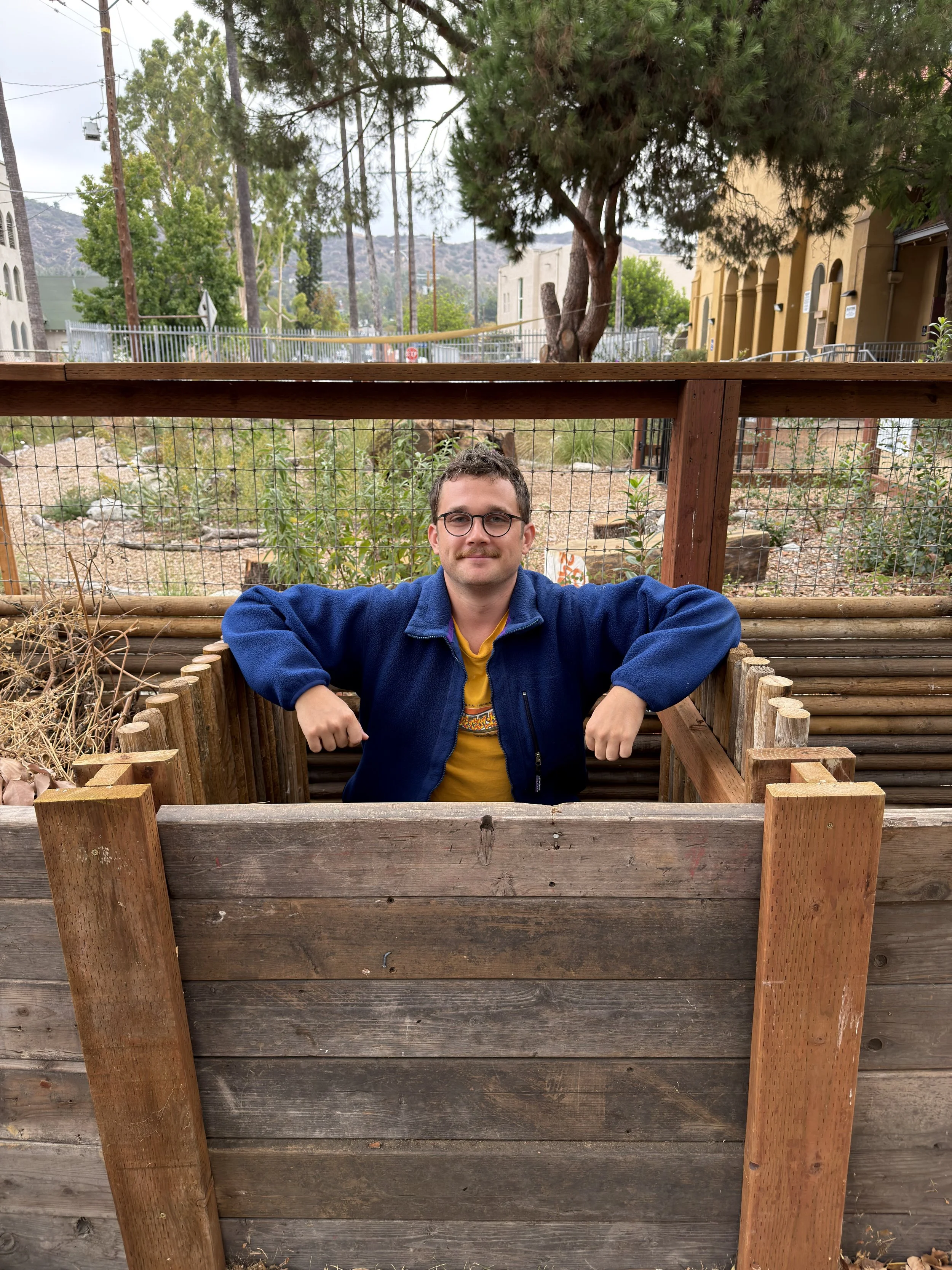 A man with glasses wearing a yellow shirt and blue jacket sitting inside a wooden compost bin, with trees and a garden in the background.