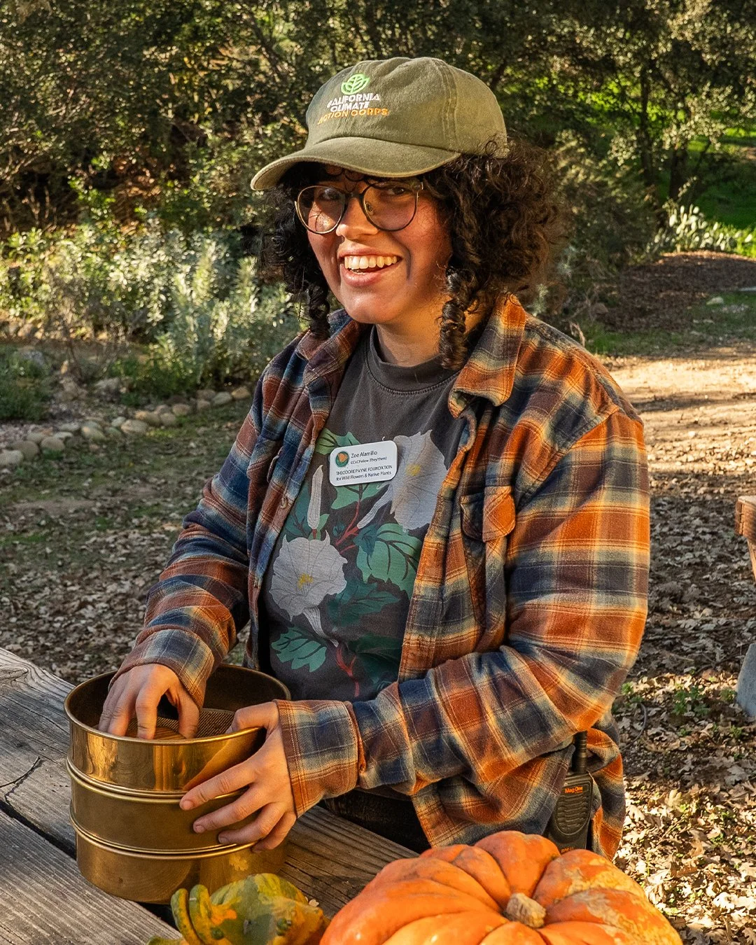 A person with curly hair, glasses, and a green cap with a logo, smiling and wearing a plaid shirt over a floral t-shirt. They are outdoors, holding a golden tin box, standing at a rustic wooden table surrounded by pumpkins and gourds.