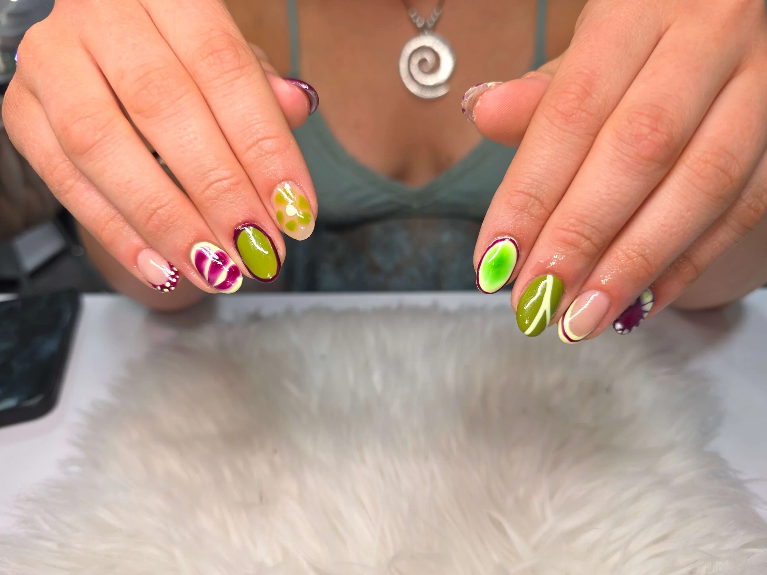 Close-up of a person's hands with colorful and decorated nail art, featuring green, pink, and white designs, resting against a blurry background.