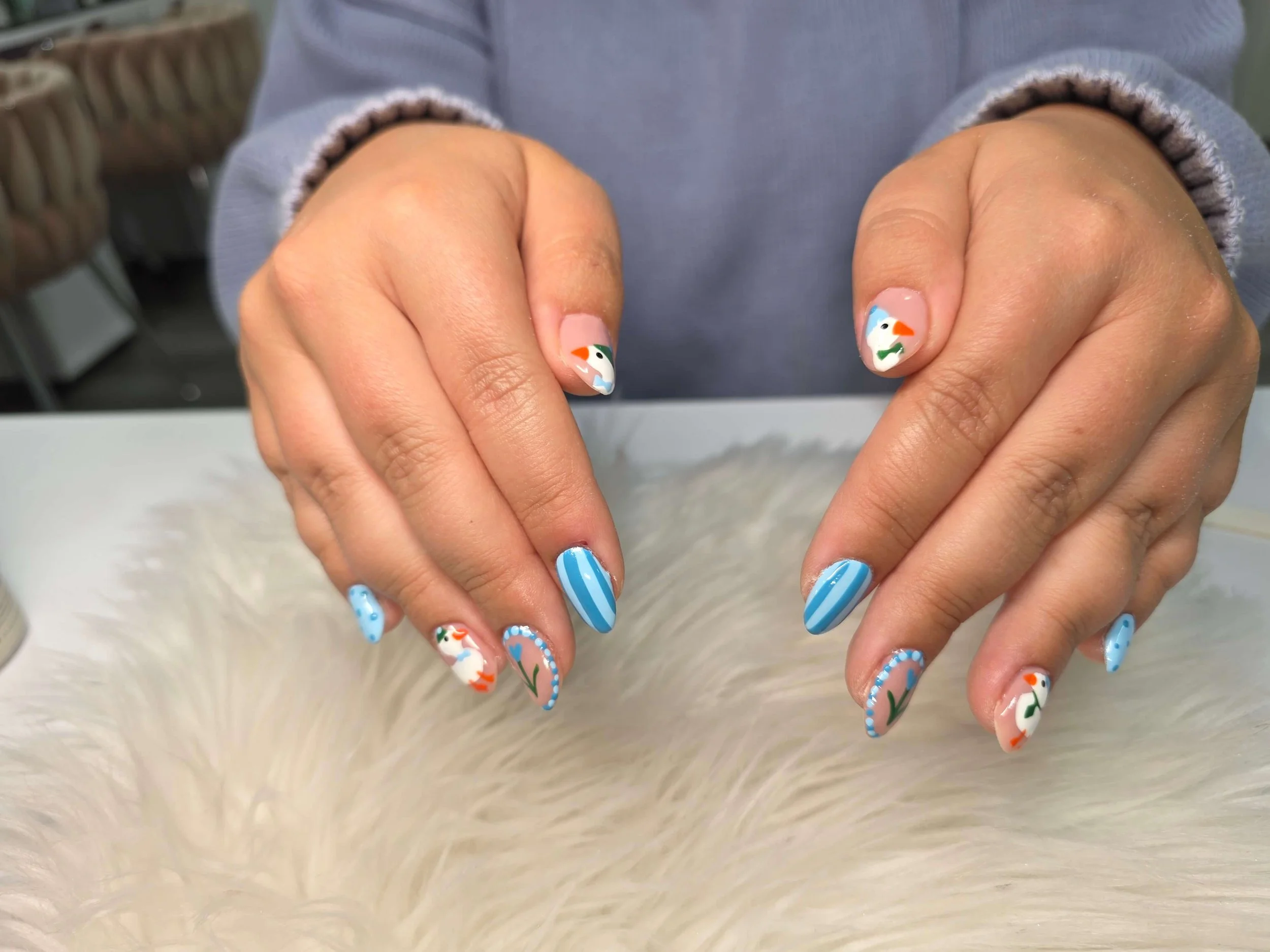 Close-up of hands with colorful, patterned nail art, featuring snowmen and stripes, resting on a fluffy white surface. A person wearing a grey sweater is in the background.